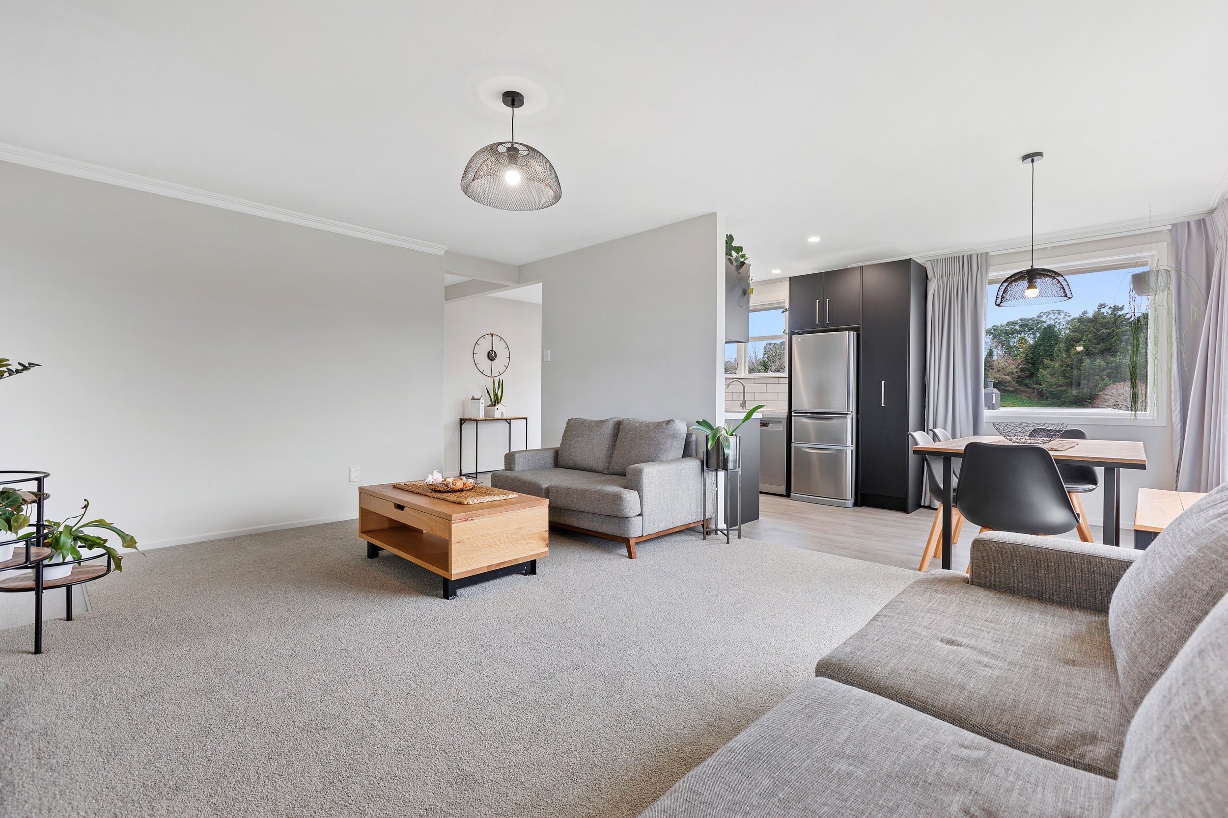 Modern living room with gray sofas, wooden coffee table, and plants, adjacent to an open kitchen with stainless steel appliances and dining area. Bright and minimalist design with neutral colors and large windows.