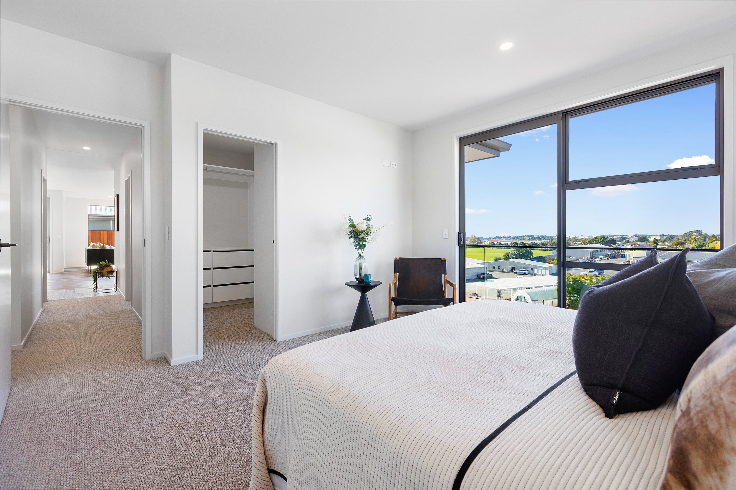 Modern bedroom with large windows, light walls, and a cozy bed. The room features a chair, side table with a vase of flowers, and a view of a green landscape outside. Adjacent hallway and closet.