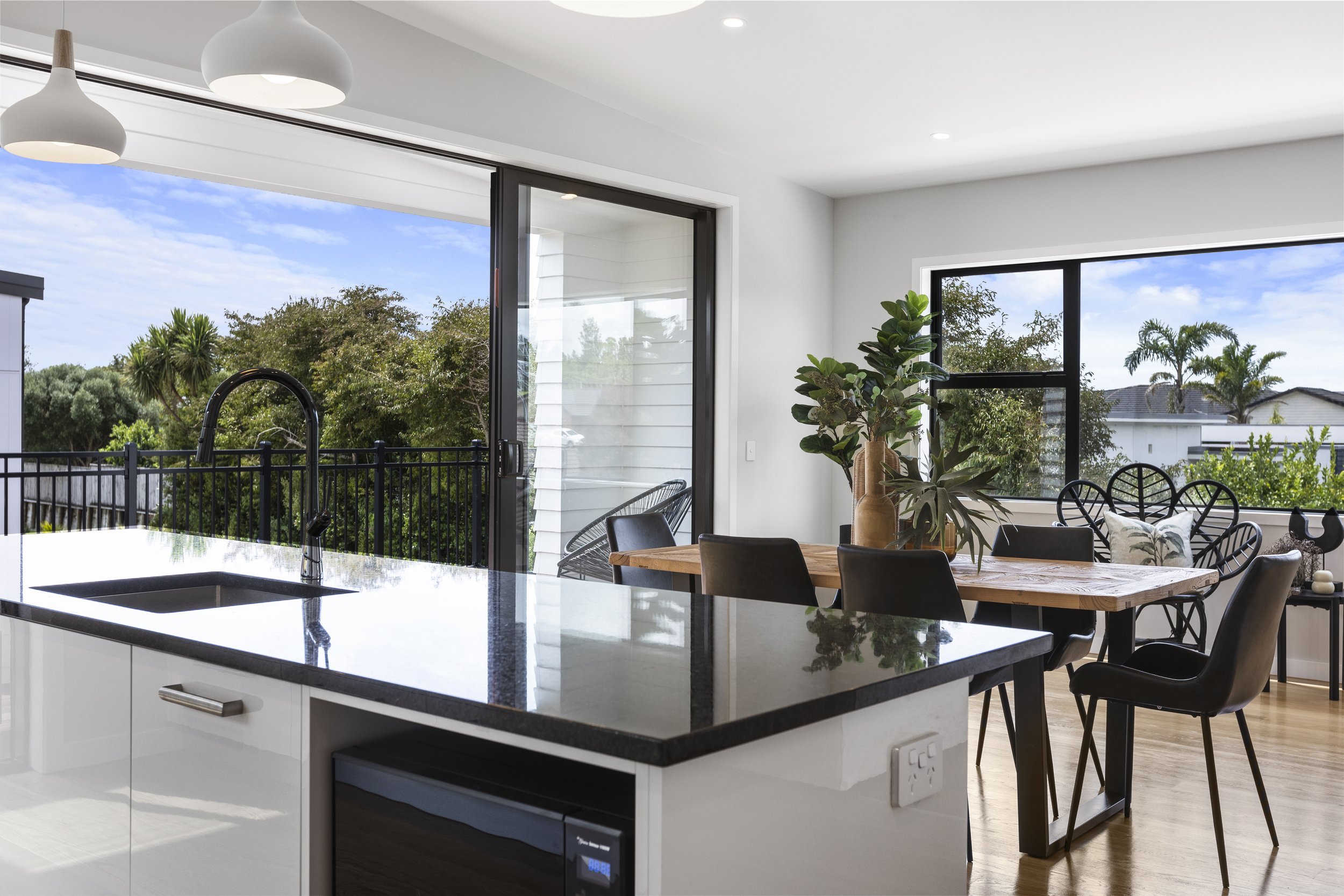 Modern kitchen and dining area with a black countertop, built-in sink, wooden dining table, black chairs, large windows, and sliding glass doors. Indoor plants and outside garden visible.