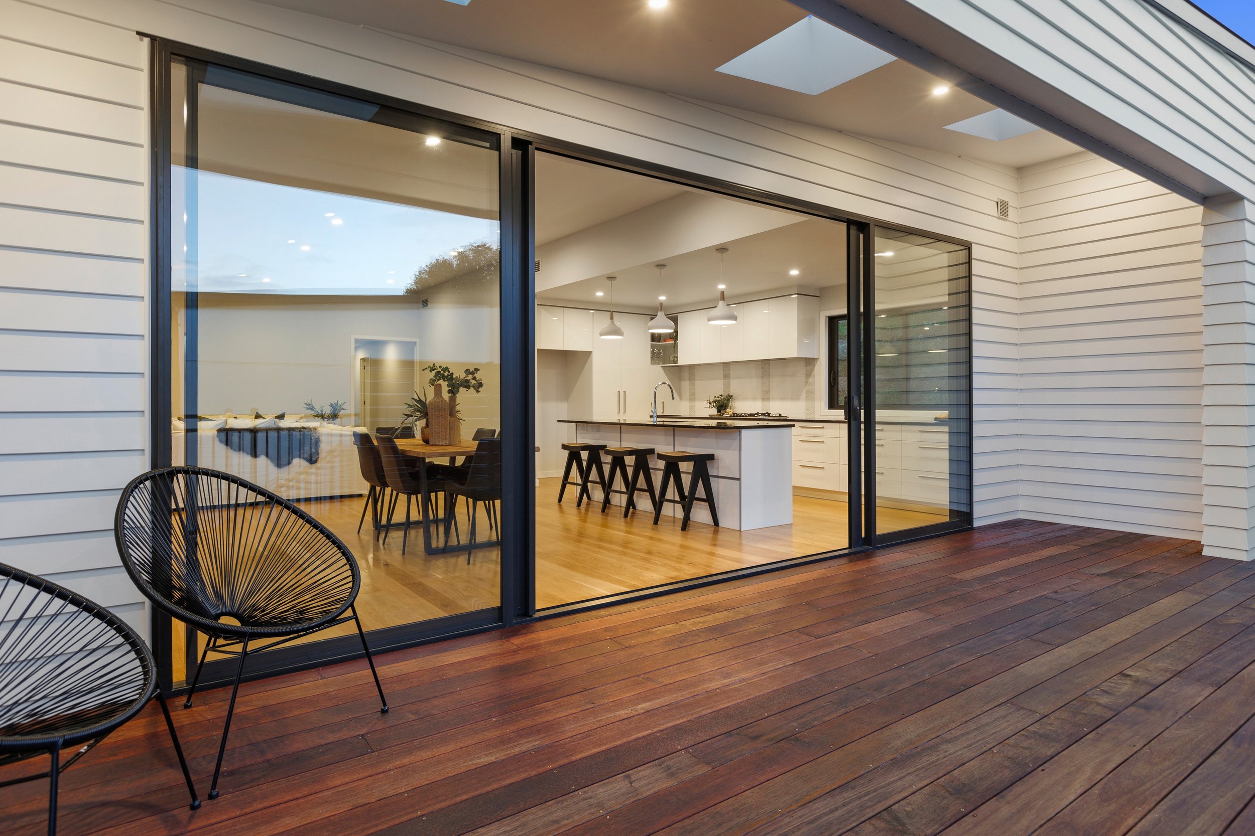 Modern patio with wooden deck, black chairs, and large sliding glass doors leading to a kitchen with an island and bar stools.