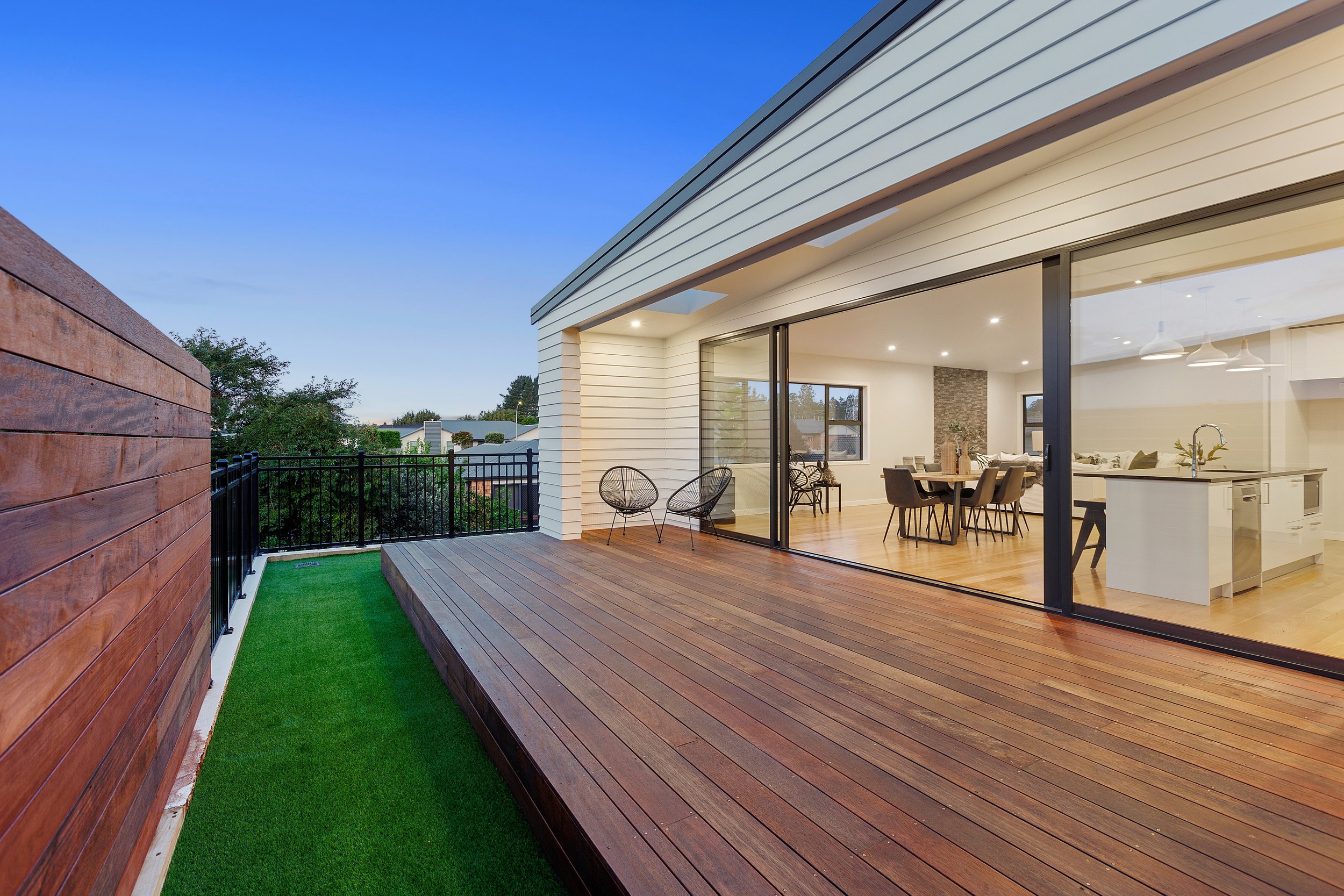 Modern outdoor wooden deck with artificial grass strip, black railings, and sliding glass doors leading to a contemporary dining and kitchen area, featuring a dining table, chairs, and pendant lighting.