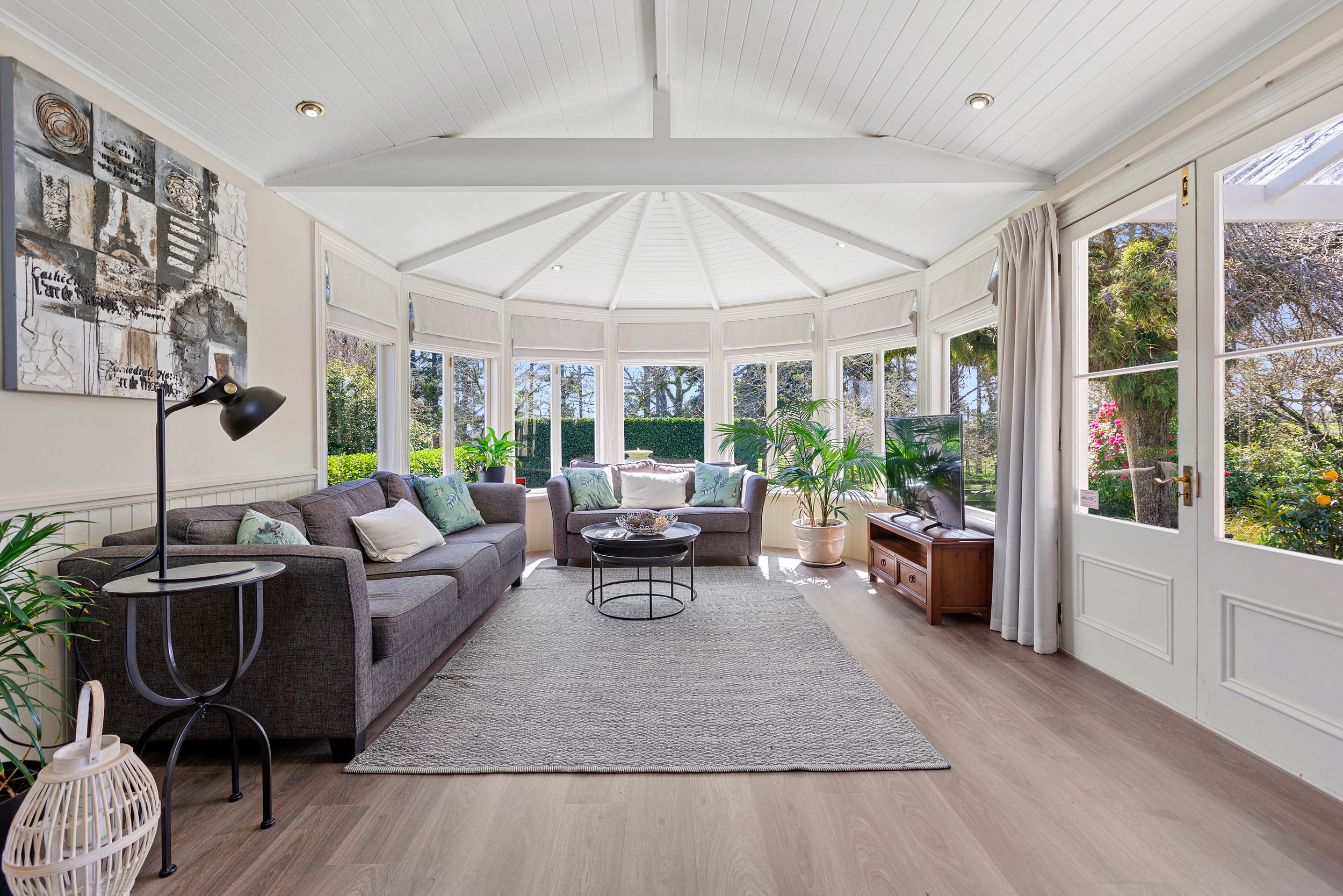Bright sunroom with large windows, gray sofas, various indoor plants, a wooden floor, coffee table, floor lamp, and abstract wall art.