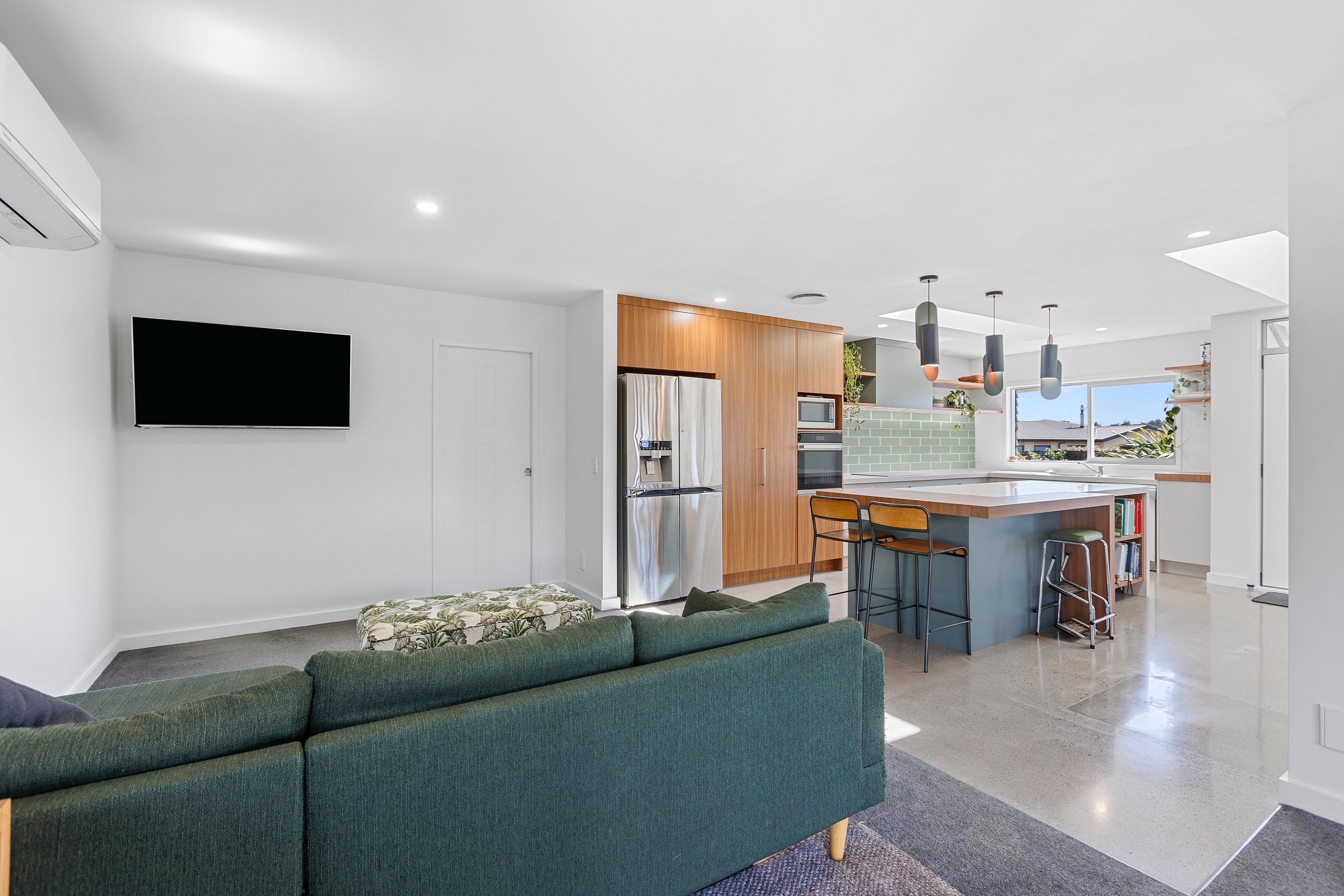 Modern open-plan kitchen and living room with green couch, wall-mounted TV, stainless steel appliances, wooden cabinets, and a kitchen island with pendant lights.