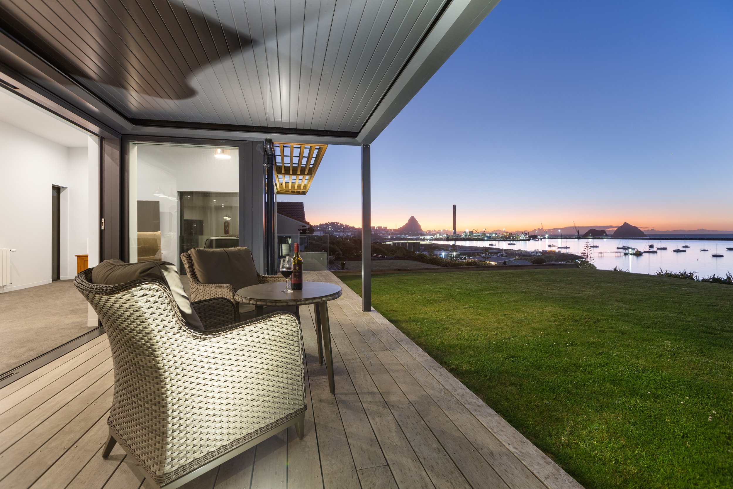 Patio with wicker chairs and table, overlooking a sunset view of a harbor with boats, grassy lawn, and distant city skyline.