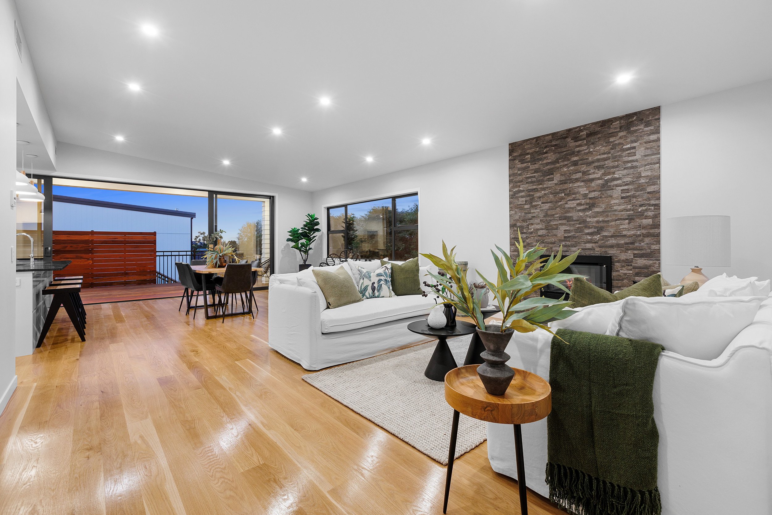 Modern living room with white sofas, wooden floors, and a brick accent wall. The space includes a dining area with a table and chairs near large windows, providing natural light. Indoor plants and minimalistic decor create a cozy ambiance.