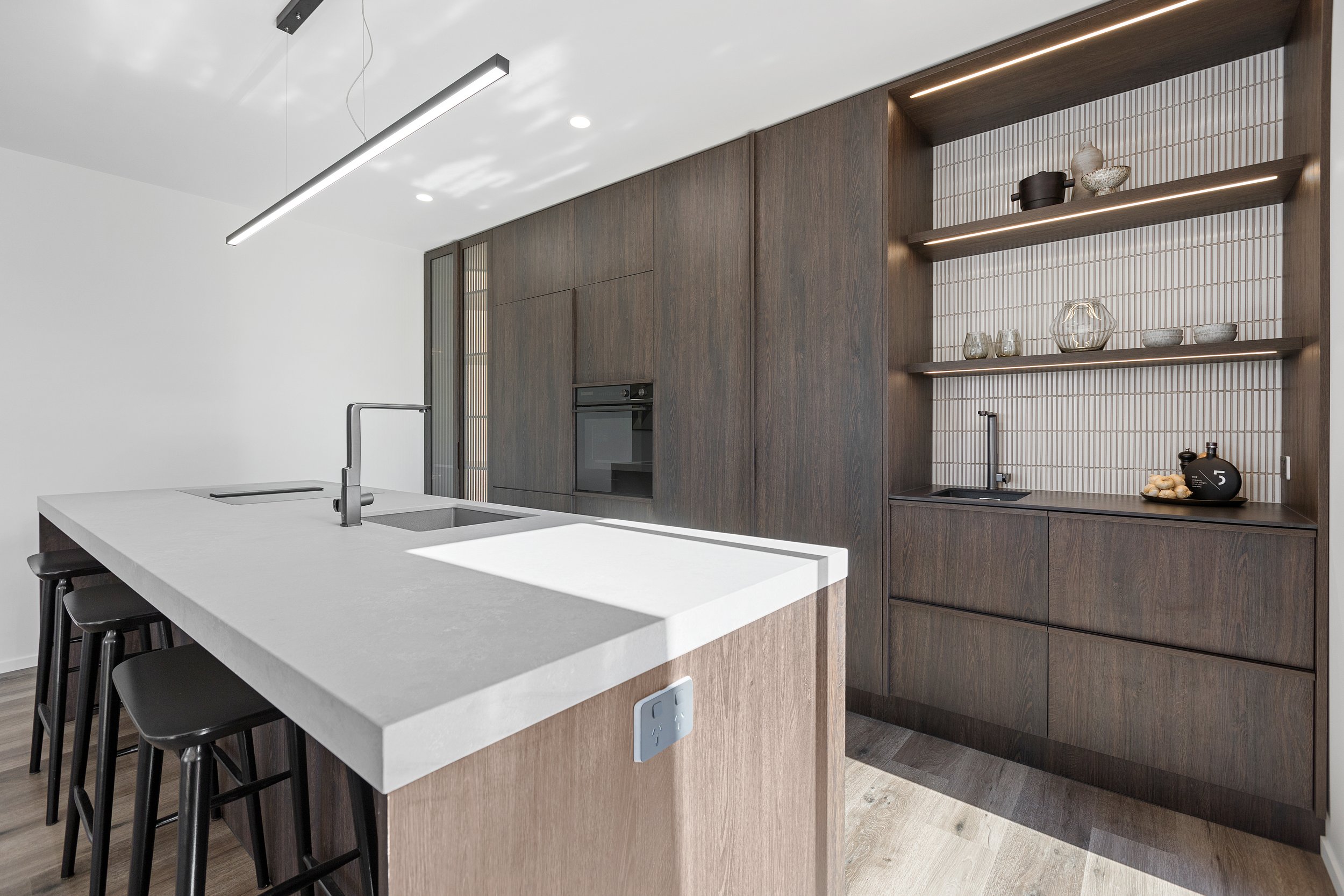 Modern kitchen interior with a large island, sleek countertop, built-in appliances, wooden cabinetry, open shelving, and contemporary lighting.