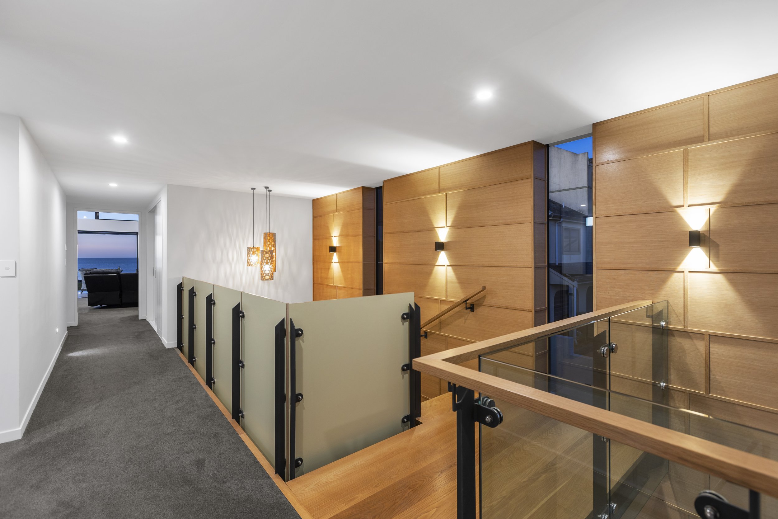 Modern interior hallway with wooden panel walls, frosted glass railing, decorative pendant lights, and a distant view of the ocean through a window.