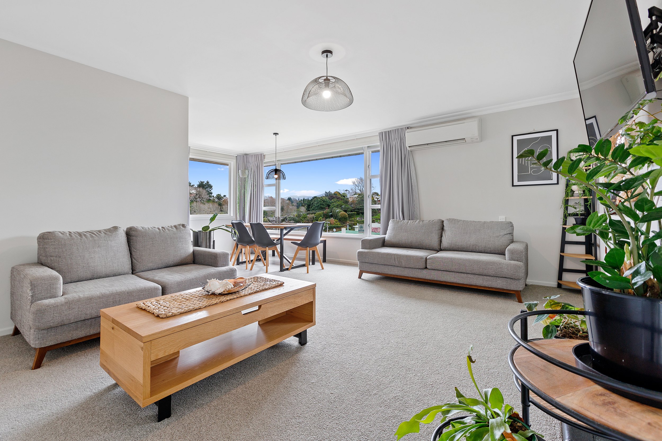 Modern living room with gray sofas, wooden coffee table, dining table with chairs by large windows, air conditioning unit, potted plants, and wall art.