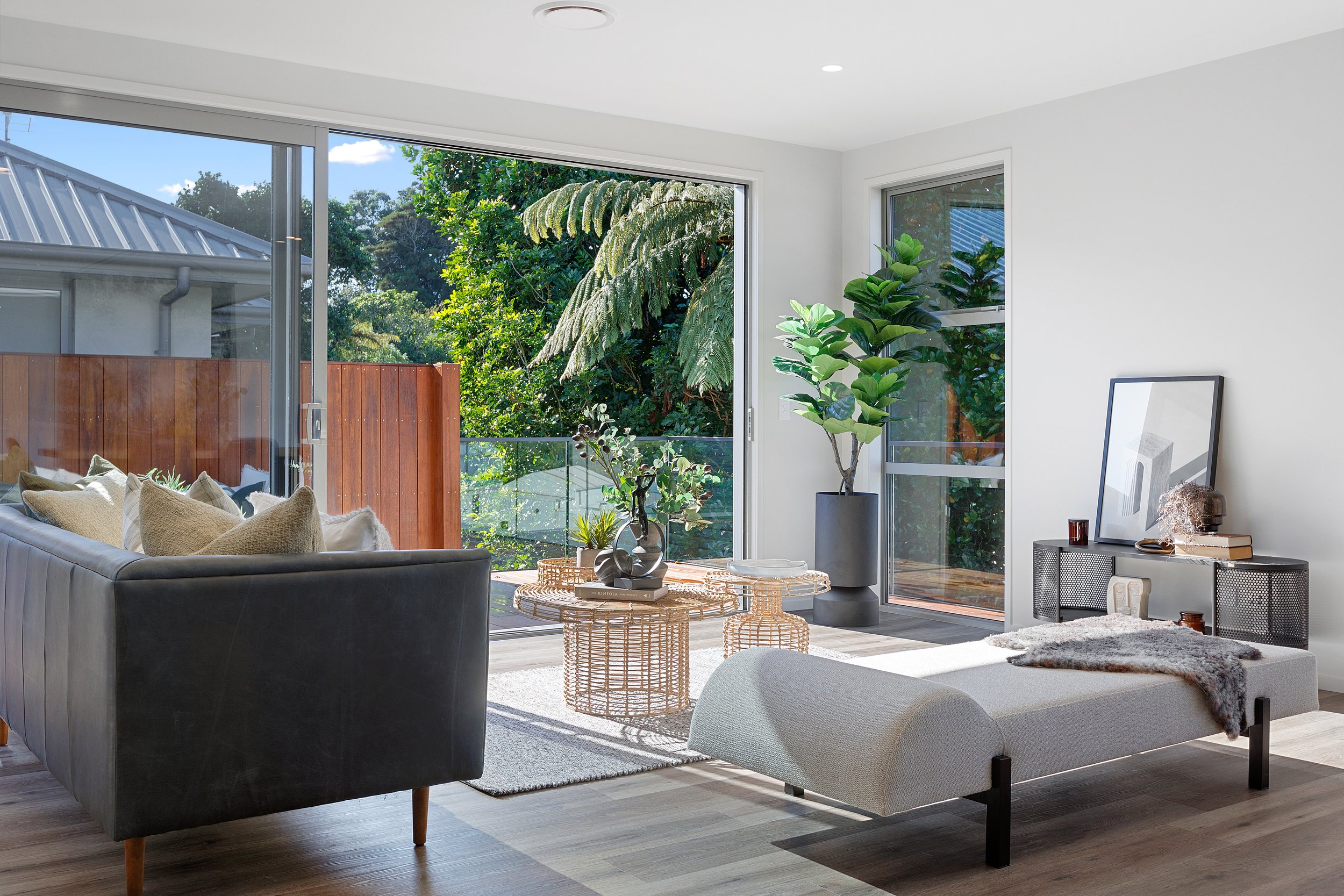 Modern living room with large windows, green sofa, wicker coffee table, potted plants, and outdoor balcony view.