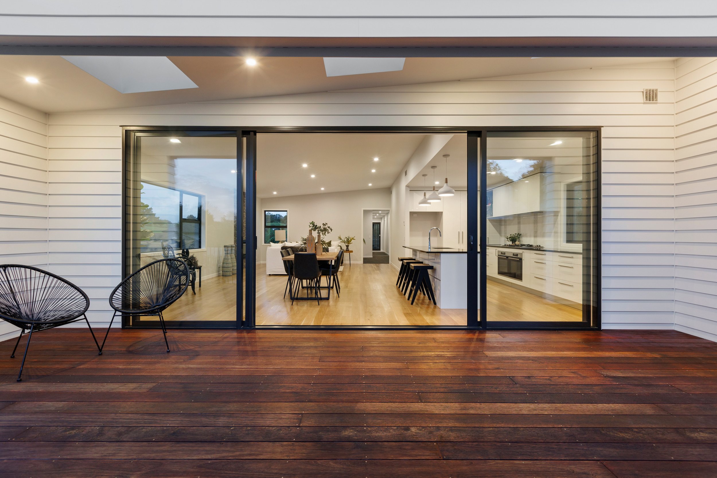 A modern open-plan interior with a view from a wooden deck. The space features a dining area and kitchen with pendant lights, black chairs, a wooden table, and bar stools. The exterior has sliding glass doors and two wireframe outdoor chairs.