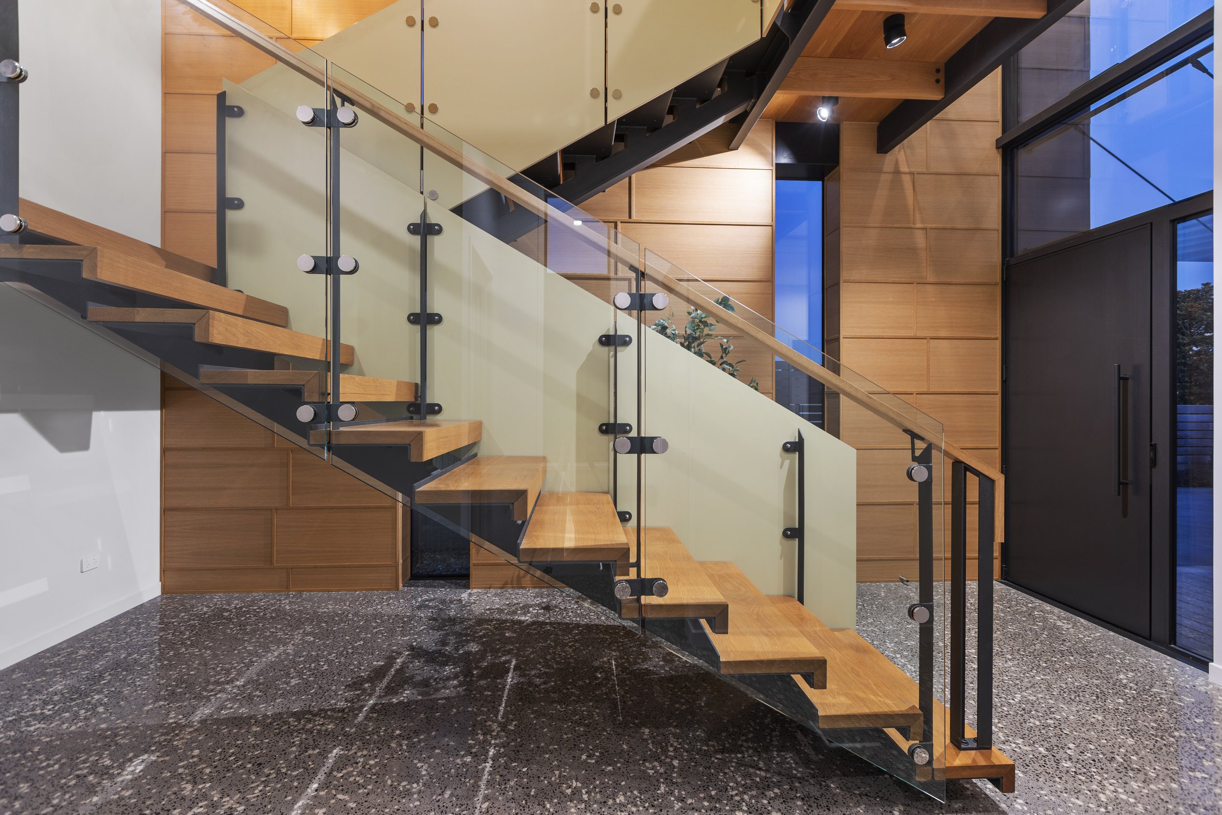 Modern staircase with wooden steps, glass panels, and sleek metal railings in a contemporary interior with wooden wall paneling and polished stone flooring.