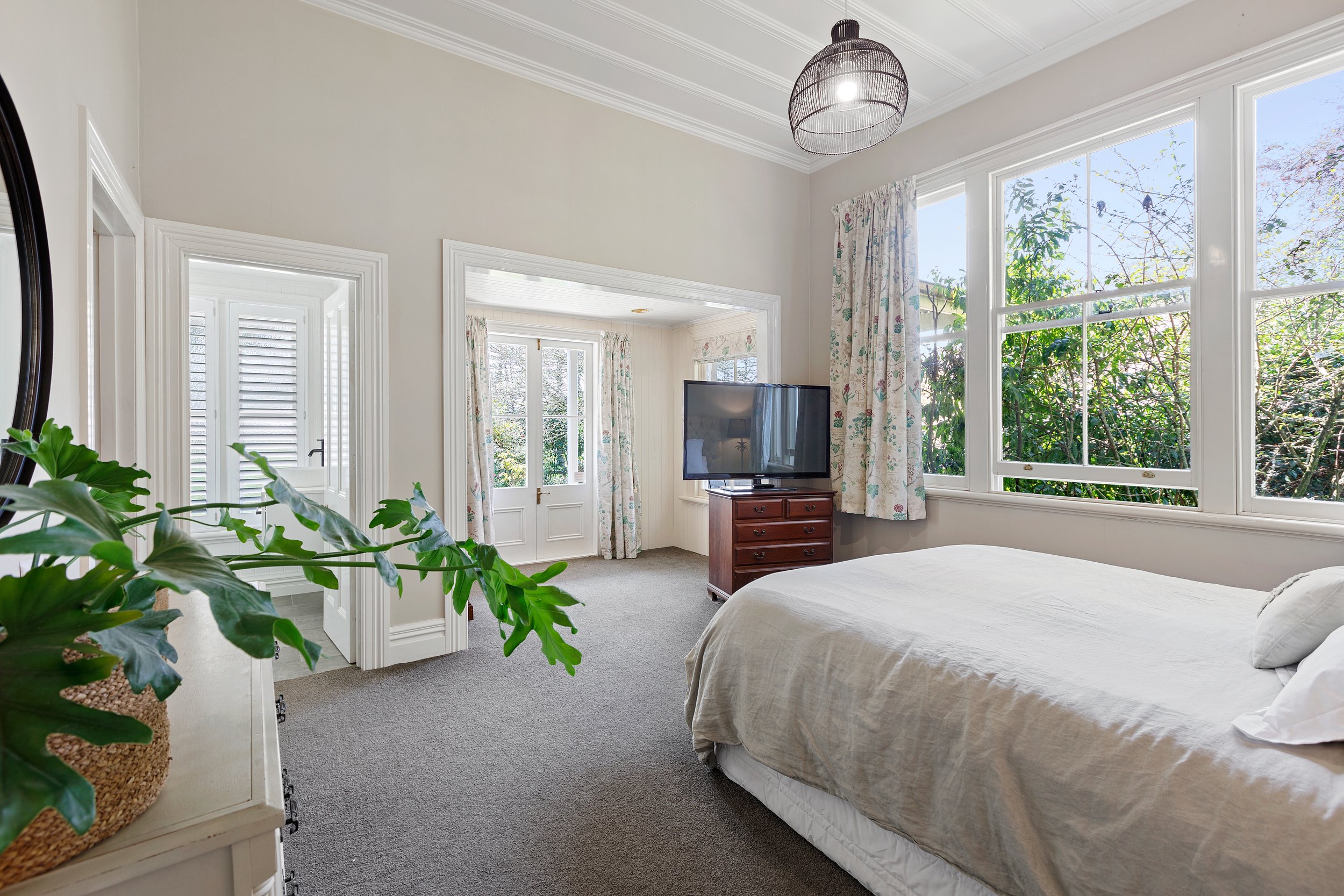 Bright bedroom with white walls, large windows with floral curtains, a double bed, a wooden dresser with a flat-screen TV, and a leafy plant.