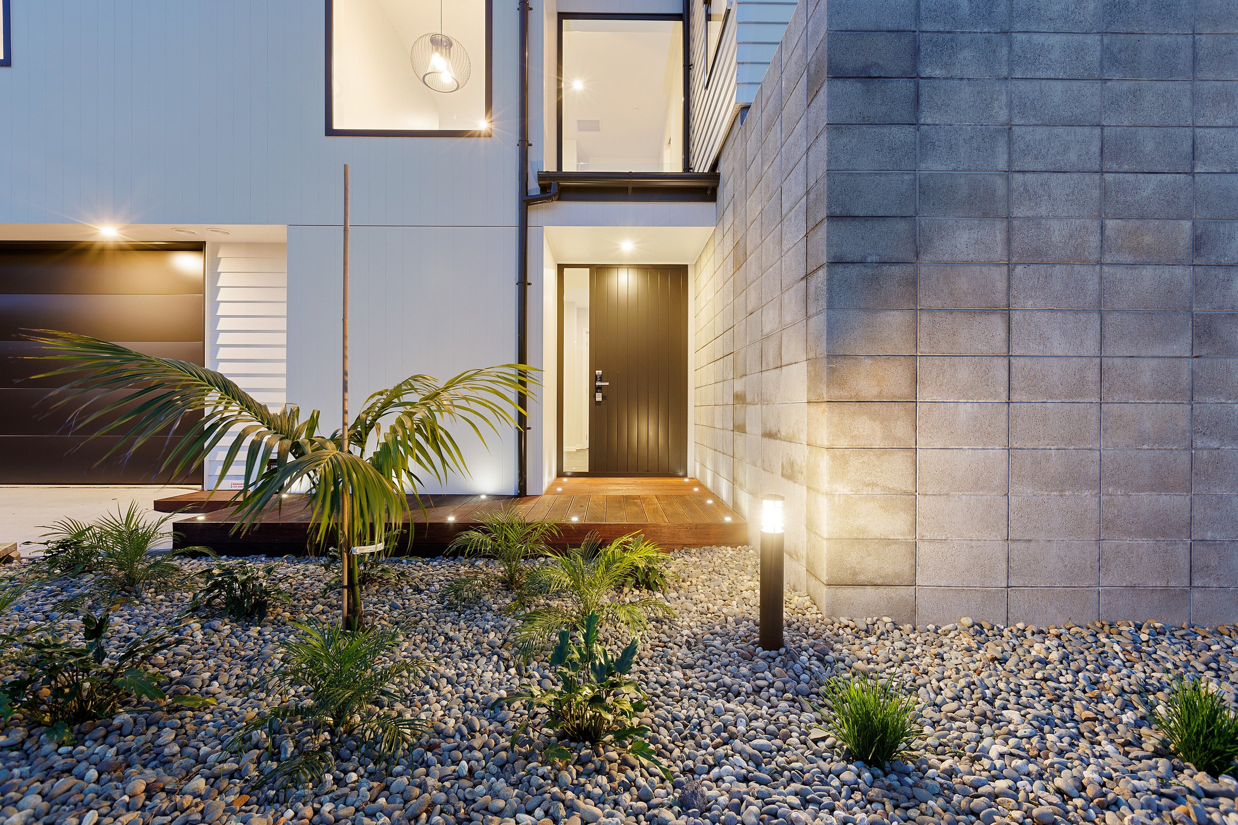 Modern house entrance with a wooden deck, large gray brick wall, and landscape lighting. The front yard features decorative gravel and tropical plants.