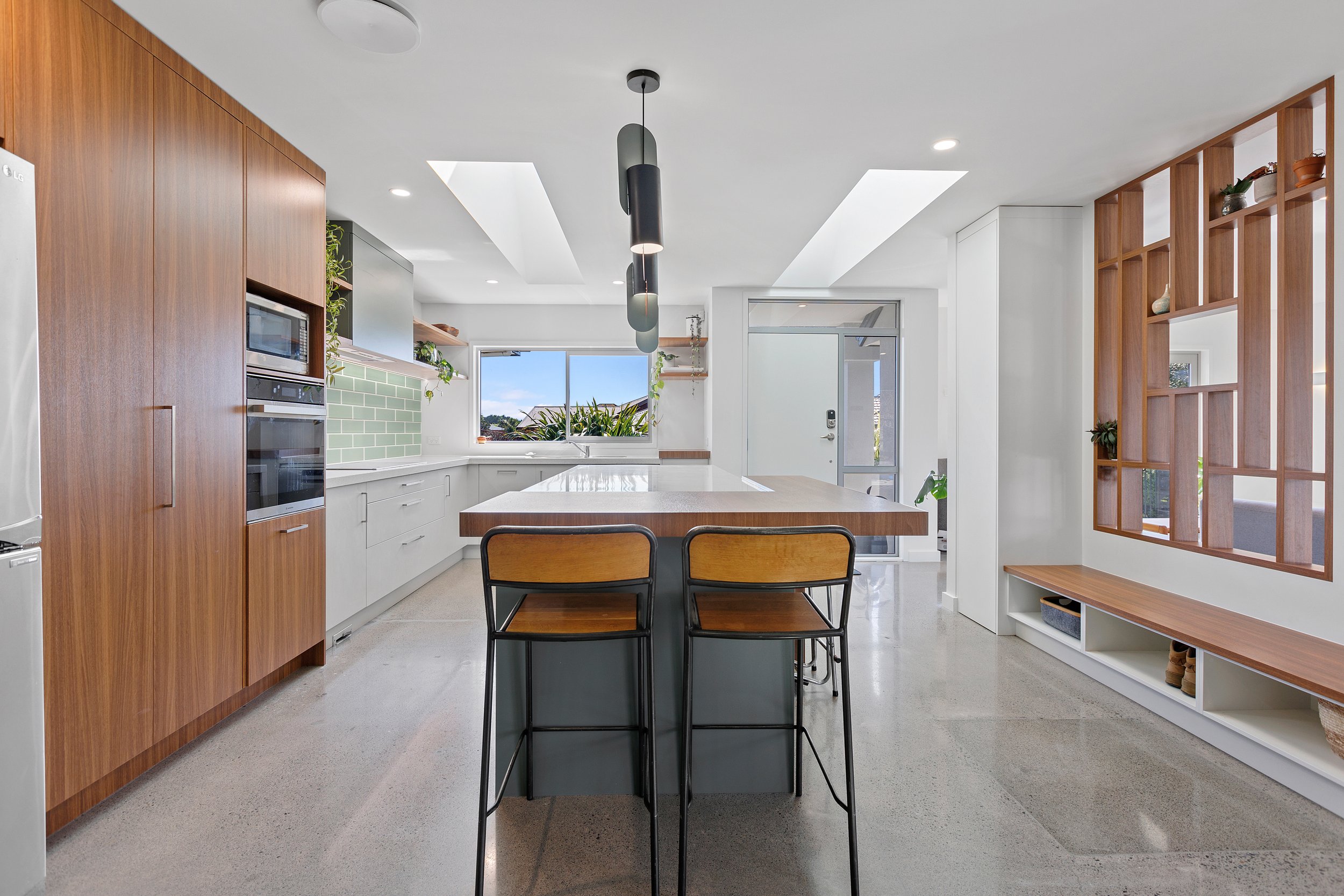 Modern kitchen with wooden cabinets, central island with bar stools, skylights, green tiled backsplash, and open shelving with plants.