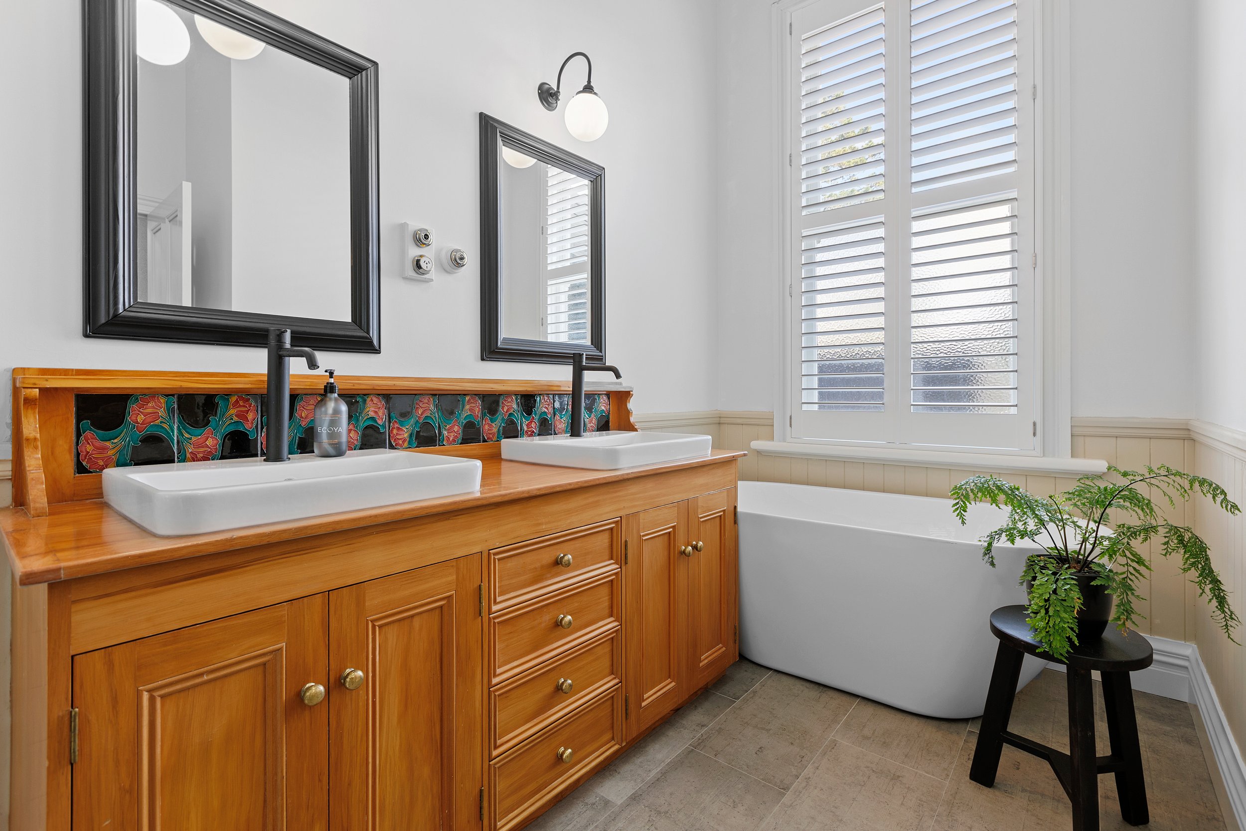 Bathroom with double vanity, wooden cabinet, two mirrors, decorative backsplash, white freestanding bathtub, window shutters, stool with plant, and tiled floor.