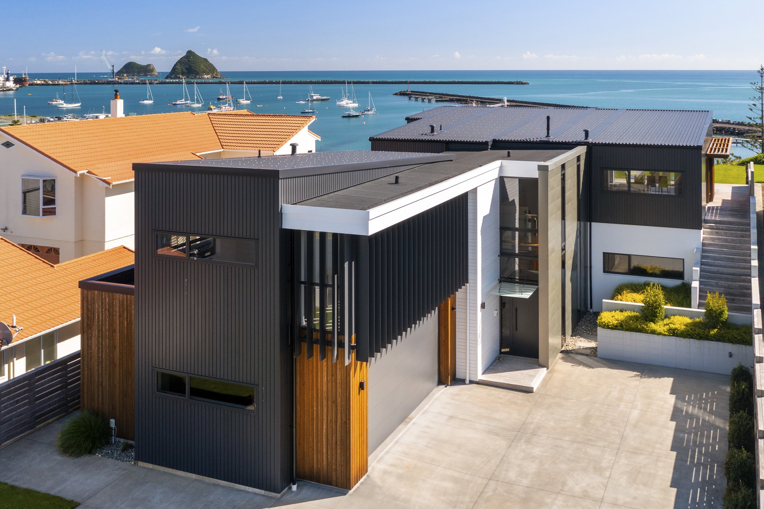 Modern coastal house with a flat roof, black and wood-paneled exterior, and large windows. The property overlooks a marina with sailboats on a calm blue sea. Adjacent homes have beige walls and orange tile roofs. A clear sky above and distant islands