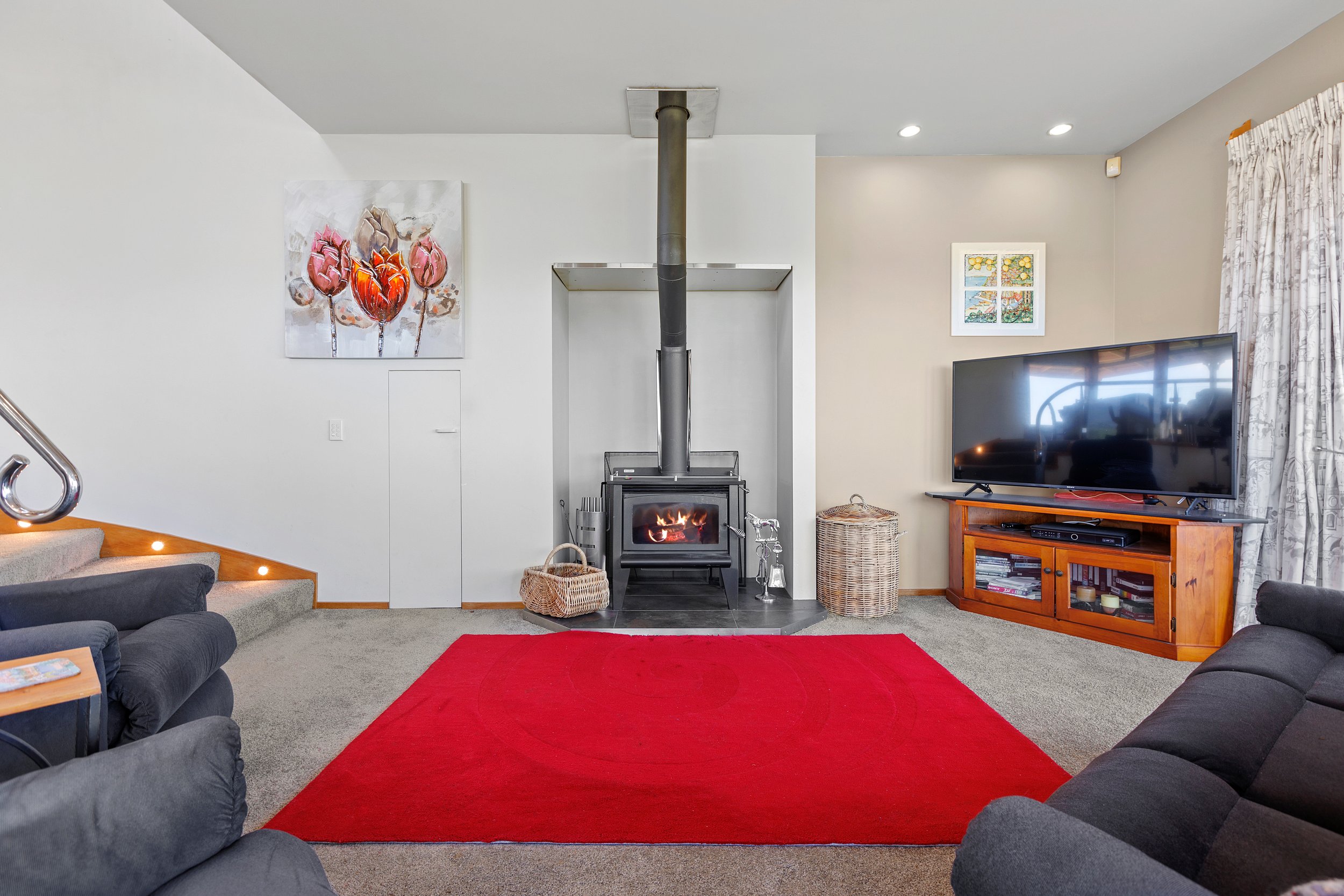 Cozy living room with a wood-burning stove, red rug, and a flat-screen TV on a wooden cabinet. A comfortable black sofa faces the stove, and a staircase with wooden steps and stainless steel railing is visible. Wall decorations include floral artwork