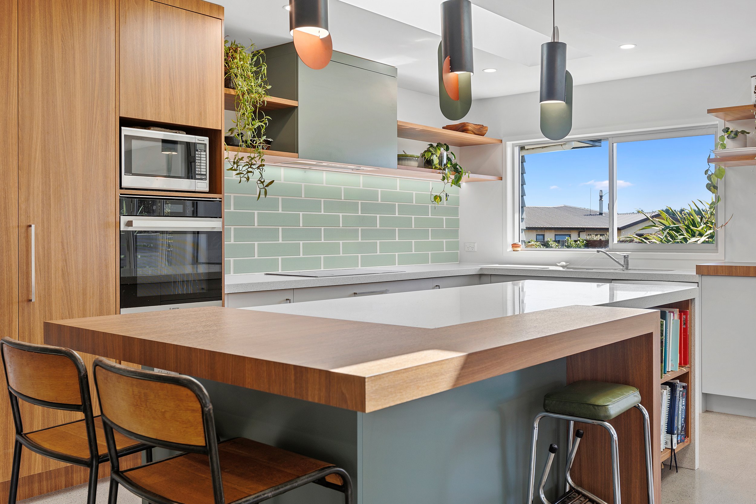 Modern kitchen interior with wood cabinetry, green subway tile backsplash, pendant lights, built-in oven and microwave, countertop seating, and large window with outdoor view.