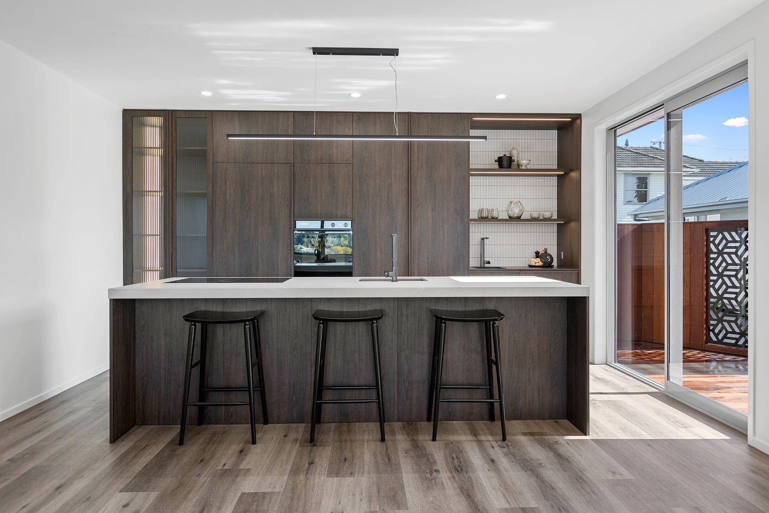 Modern kitchen with wooden cabinets and island, barstools, built-in oven, open shelving, large window with balcony view, and contemporary lighting.