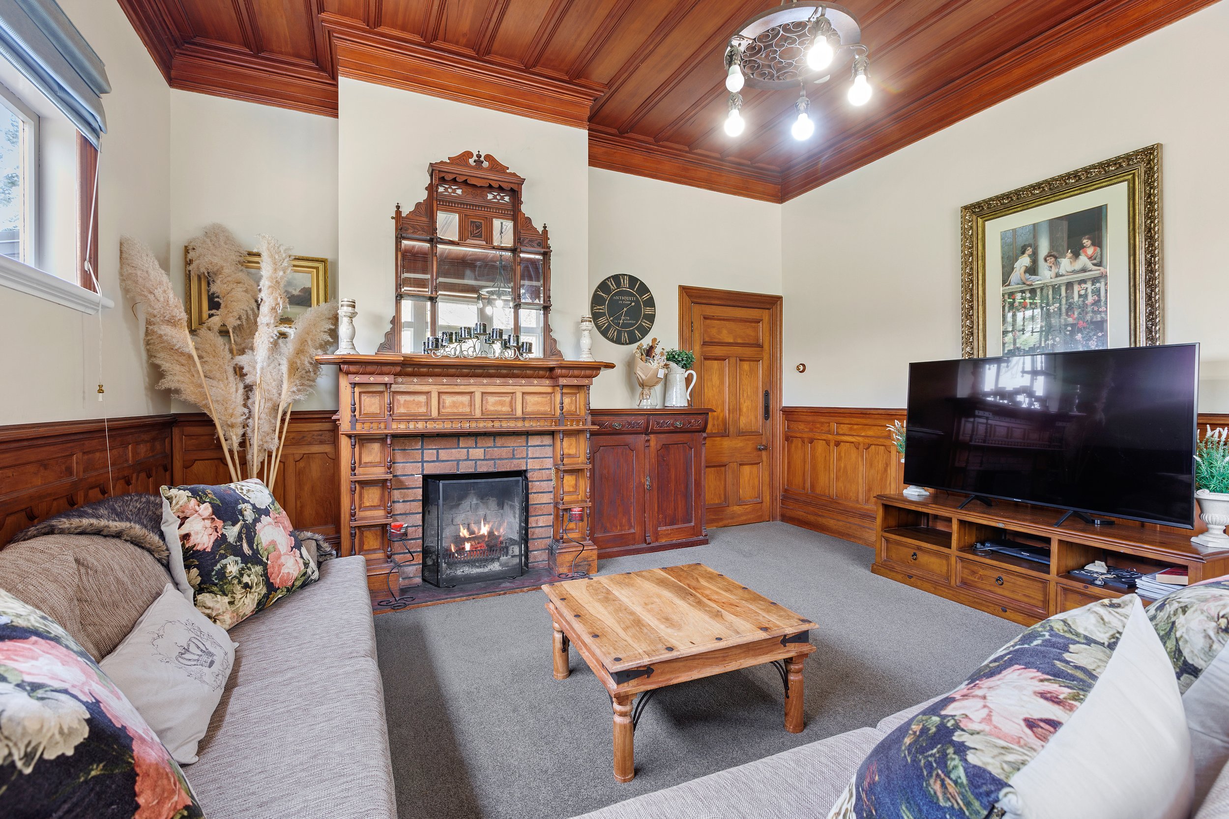 Traditional living room with wooden paneling, a large framed mirror above a fireplace, and a flat-screen TV. The room features a wooden coffee table, floral pillows on a sofa, pampas grass decor, and a vintage wall clock. A painting hangs on the wall