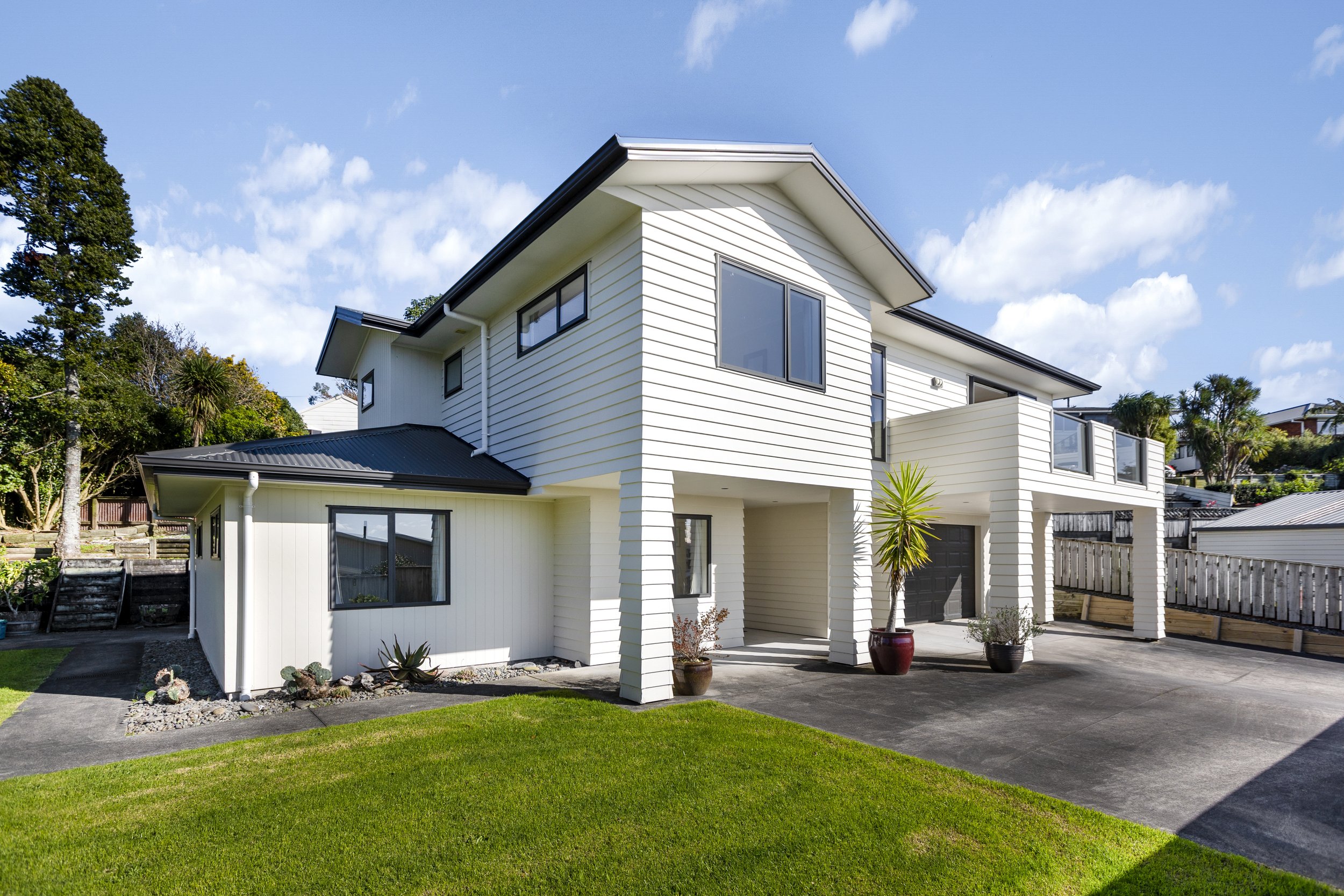 Modern two-story white house with a sloped roof, large windows, and a spacious driveway. Surrounded by a well-maintained lawn and landscaped elements, with a clear blue sky overhead.