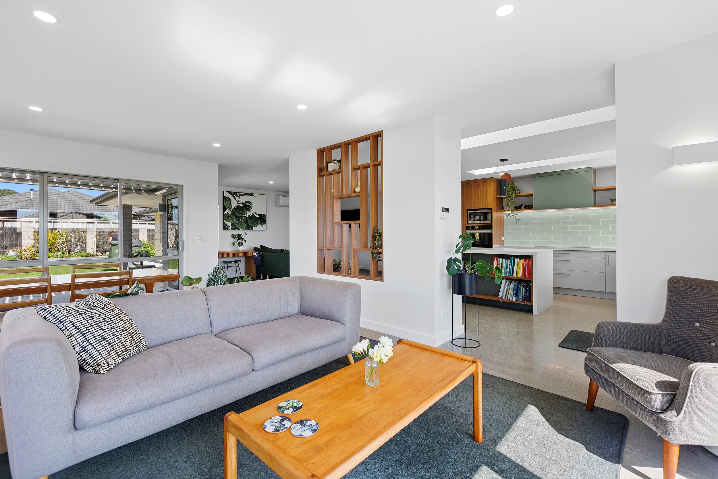 Modern living room and kitchen with gray sofa, wooden coffee table, bookshelves, and greenery. Large windows offer garden view, and kitchen has light green backsplash.