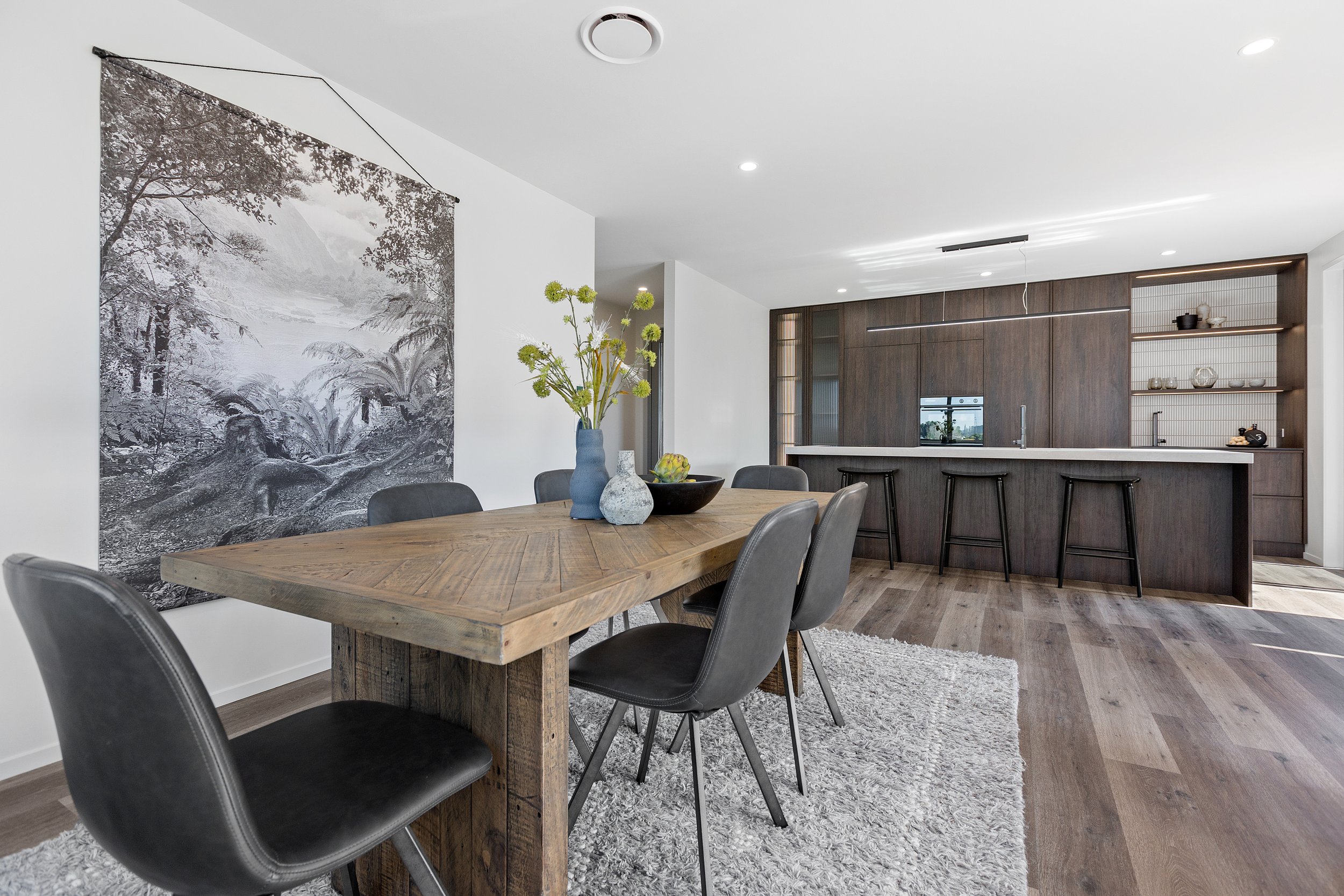 Modern dining area with wooden table, black chairs, vase of flowers, wall art, and adjacent kitchen with dark cabinetry and bar stools.