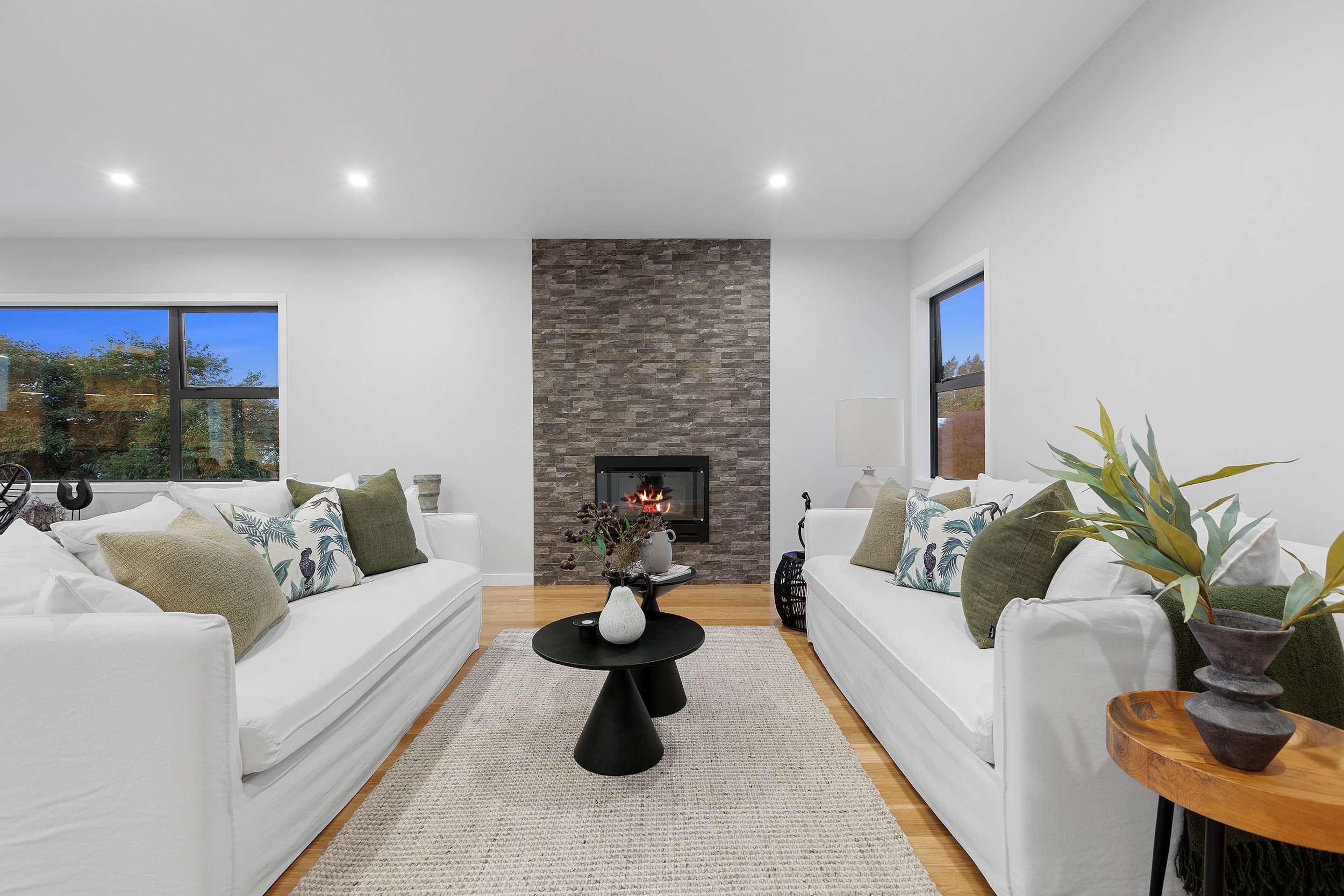 Modern living room with two white sofas facing each other, a black round coffee table, and a stone fireplace. The room features a large window, light wood flooring, and neutral-toned pillows and decor.
