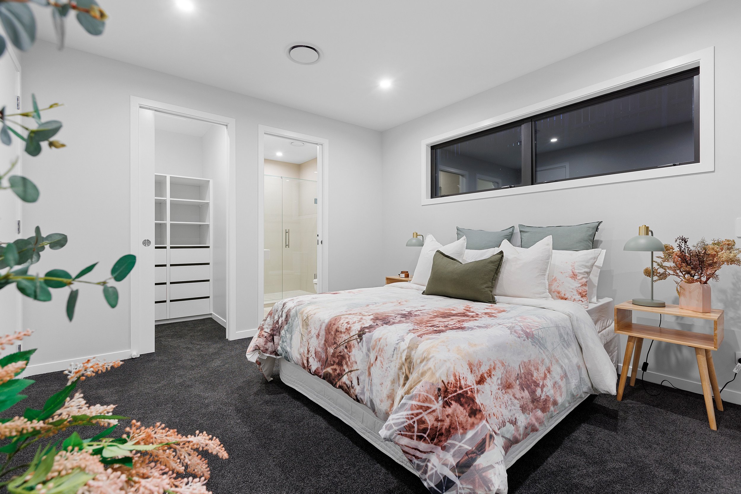 Modern bedroom interior with a bed featuring white and floral bedding, green accent pillows, and a wooden side table with a lamp and plant. The room has a walk-in closet and an ensuite bathroom visible in the background. Large window and recessed lig