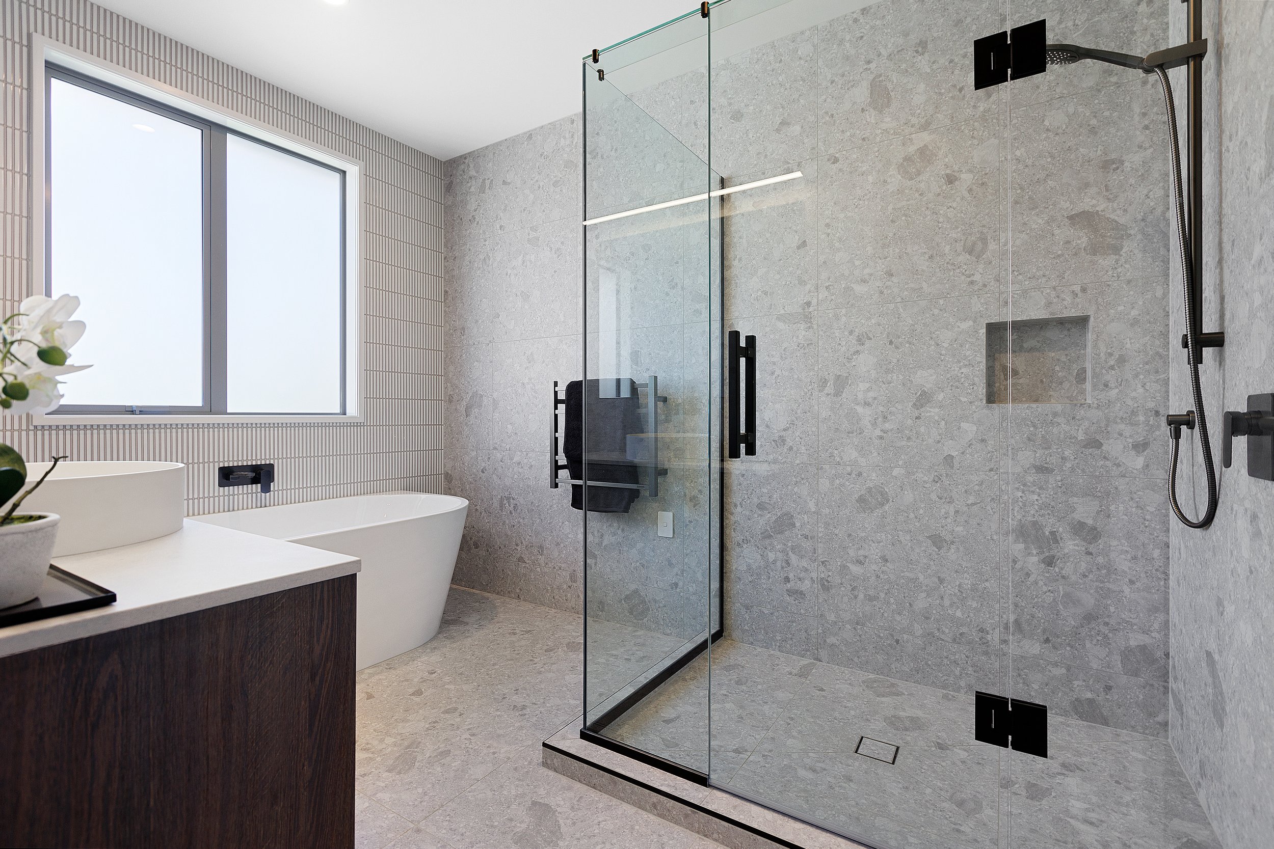 Modern bathroom with a glass shower enclosure, gray tile walls, large window, freestanding bathtub, dark wood vanity, and a white countertop with a potted orchid.