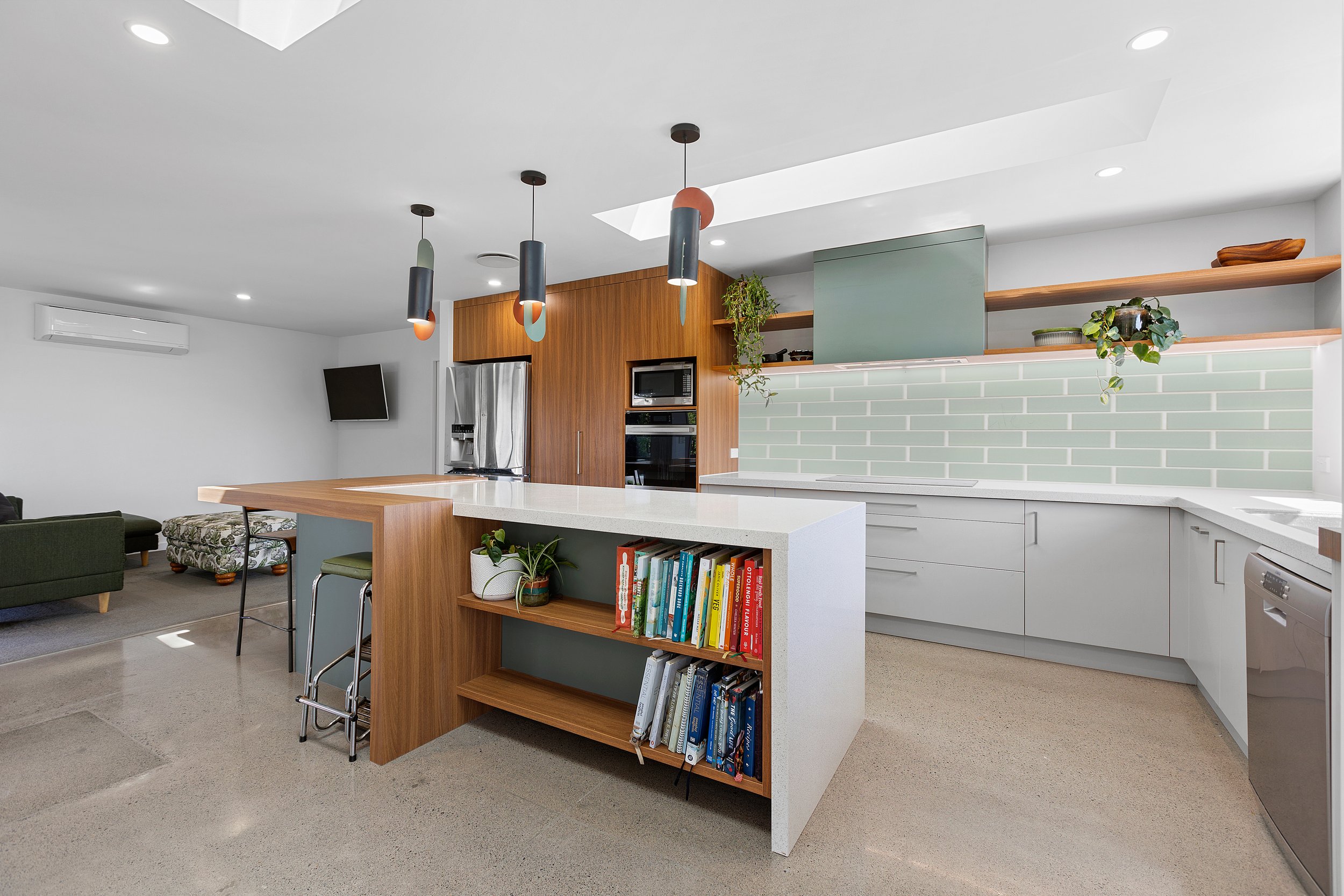 Modern kitchen with white countertops, wooden cabinets, mint green subway tile backsplash, and pendant lights. Open shelving with plants and books, stainless steel appliances, and a lounge area visible in the background.