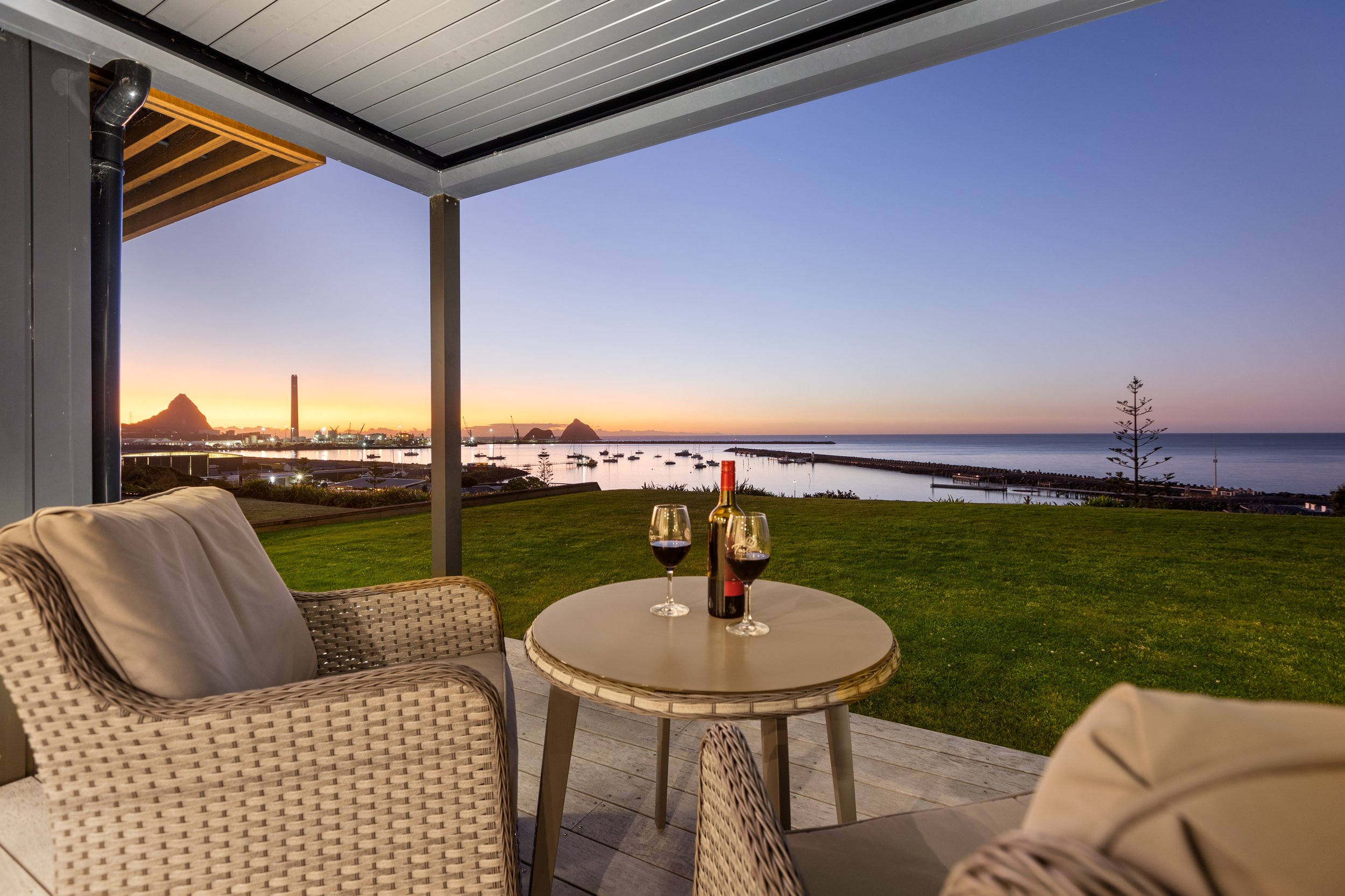 Outdoor patio with wicker chairs, table holding two wine glasses and a bottle, overlooking a sunset view of a harbor with boats and distant mountains.