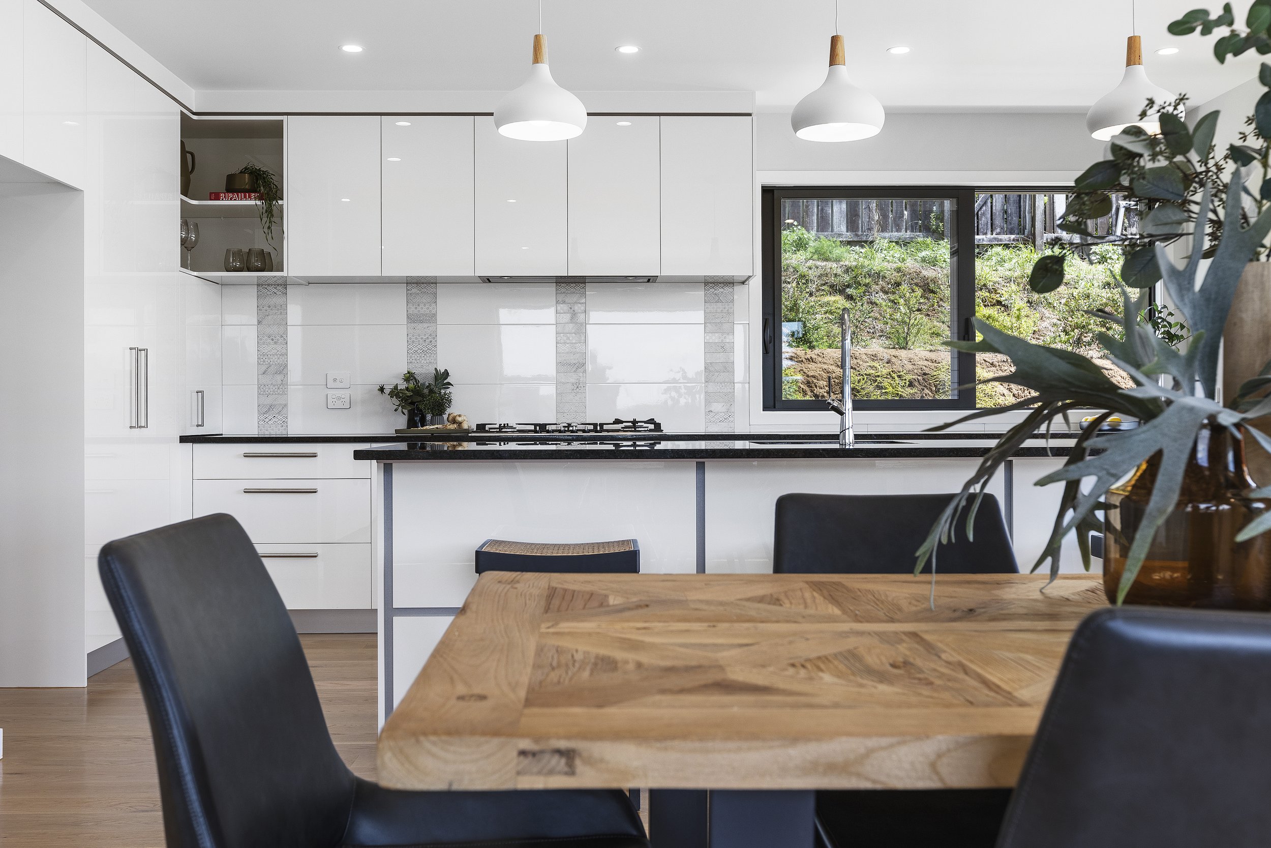 Modern kitchen with white cabinets, black countertops, and wood dining table. Pendant lights hang overhead. A large window shows greenery outside.
