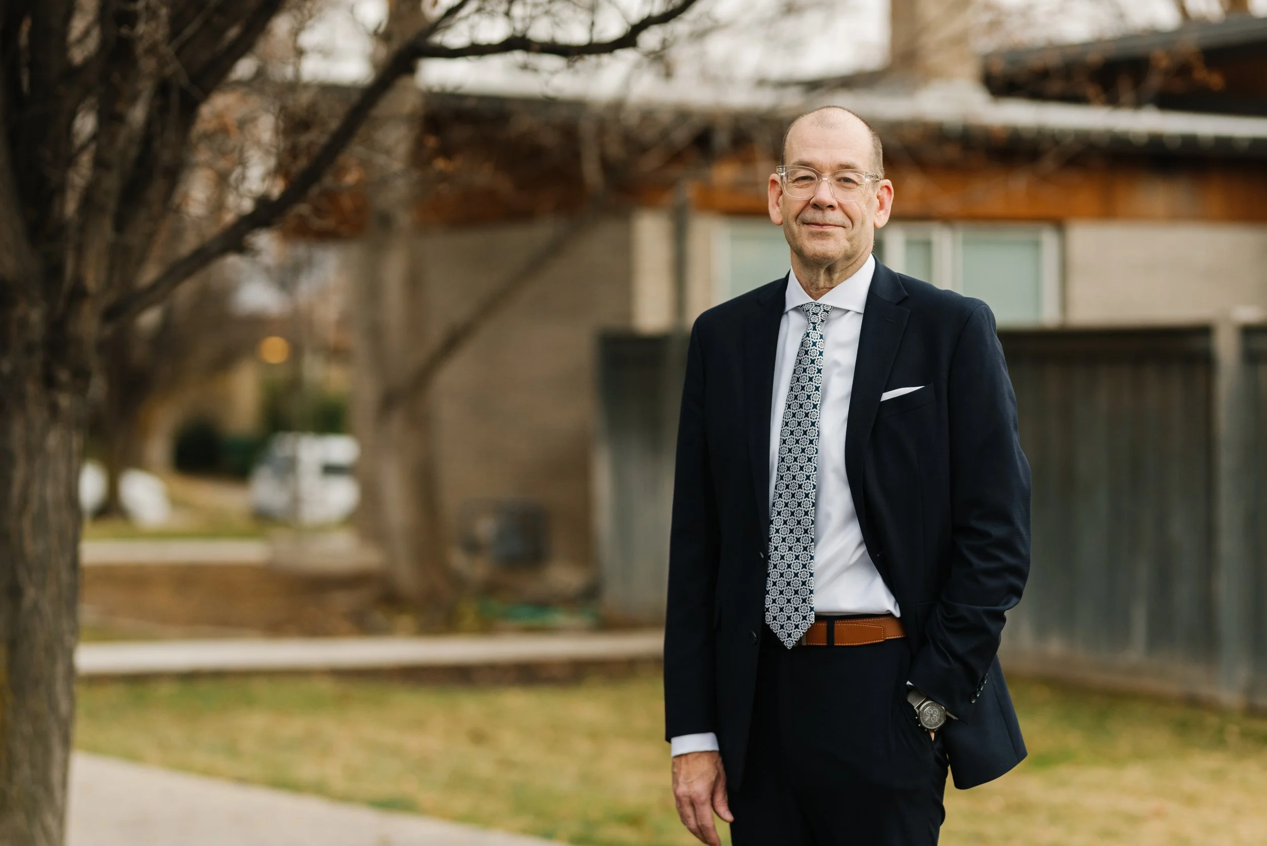 McKay Jensen in a dark suit, white shirt, and patterned tie standing outdoors near trees with bare branches, with a residential neighborhood in the background.
