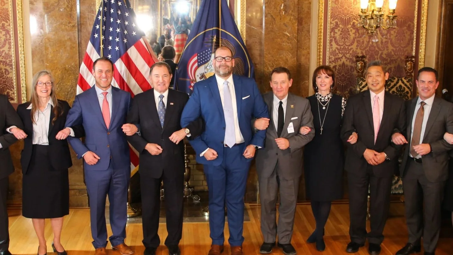 McKay Jensen with Utah leaders, posing for a photo in front of American and Utah state flags. They are dressed in formal business attire.