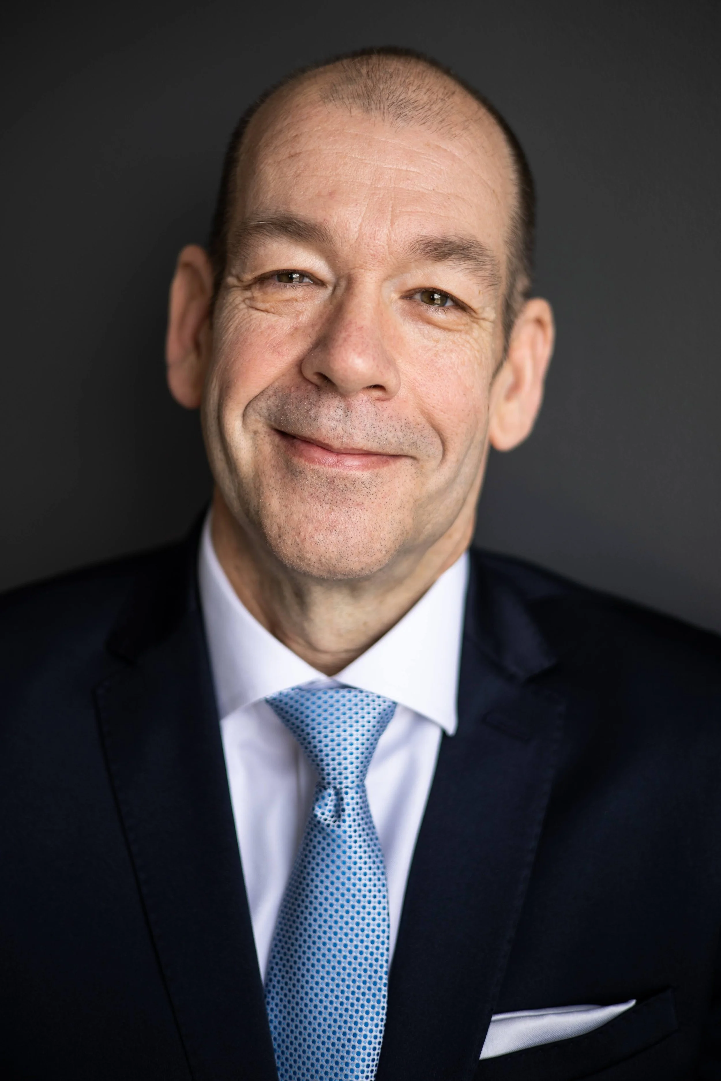 A McKay Jensen in a dark suit, white shirt, and blue tie smiling against a dark background.