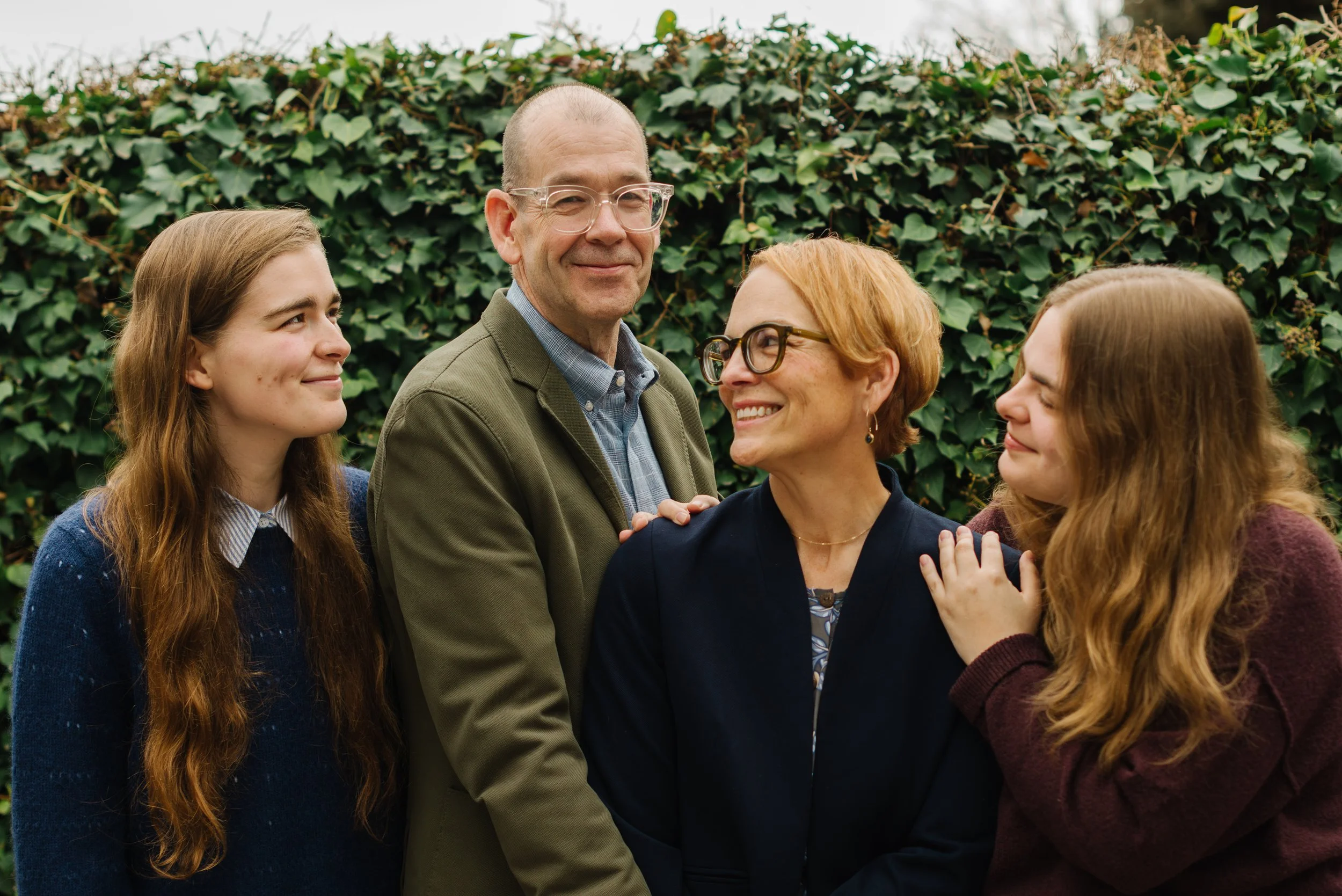 A family of four standing outdoors in front of green ivy, smiling and looking at each other.