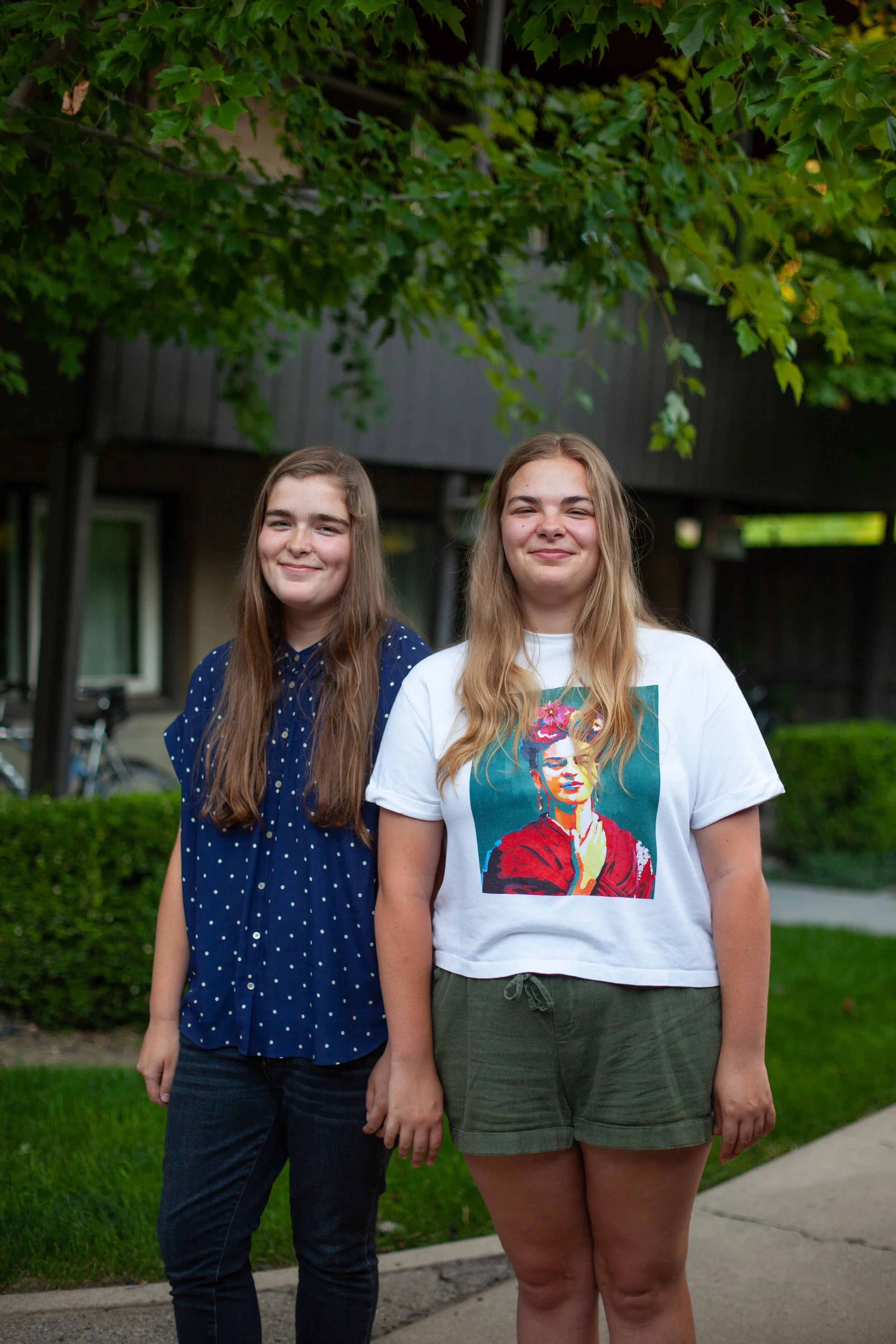 Lauren and Lily standing outdoors on a sidewalk with green bushes and a building in the background, smiling at the camera.
