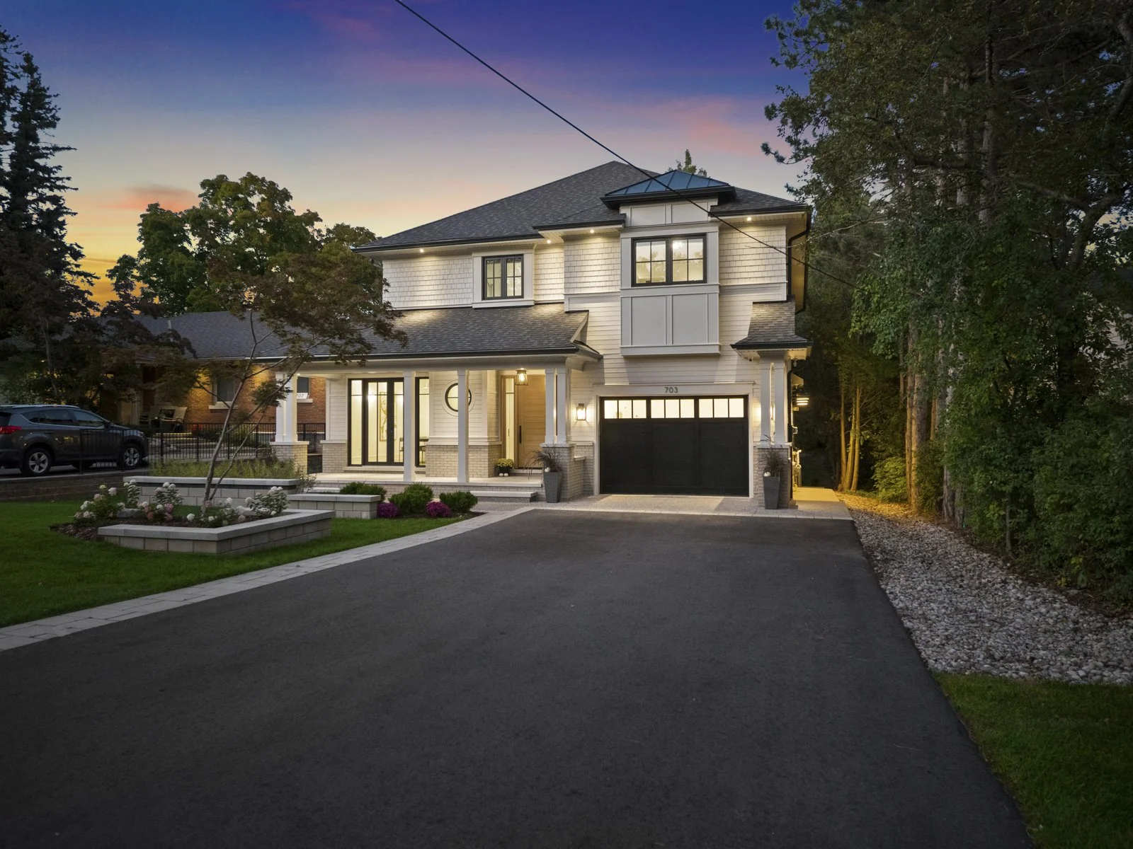 A modern two-story custom built house illuminated at dusk, with a black garage door, white exterior, and landscaped front yard featuring flower beds and a paved driveway.