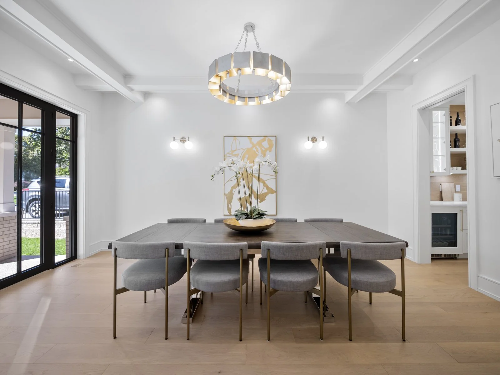 Modern dining room with a large wooden table and ten gray chairs, a decorative orchid centerpiece, white walls with artwork, a large circular chandelier, and sliding glass doors leading outside.