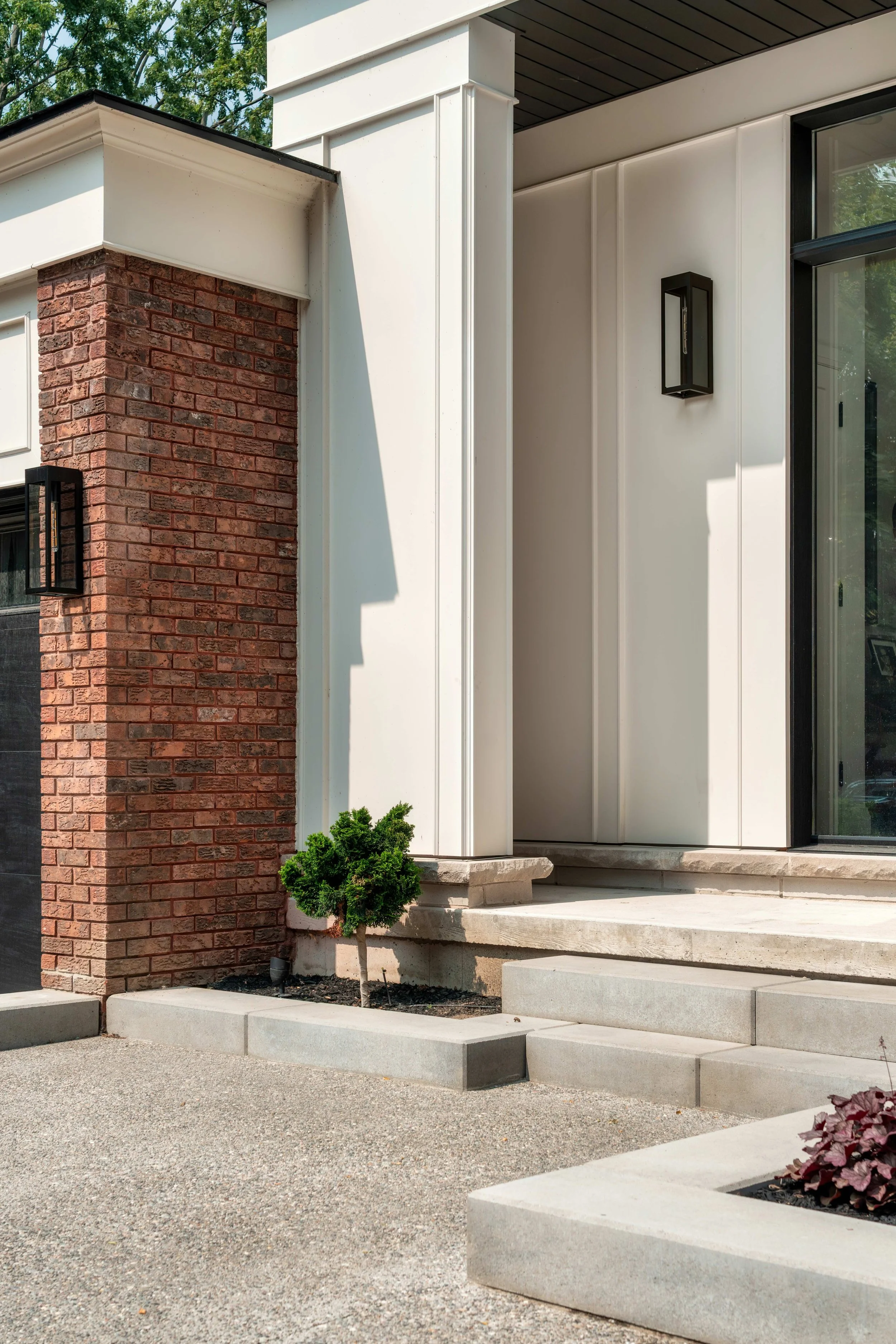 Modern residential exterior featuring a small tree in a garden bed, concrete steps, and a brick and white siding facade with black outdoor wall lights.
