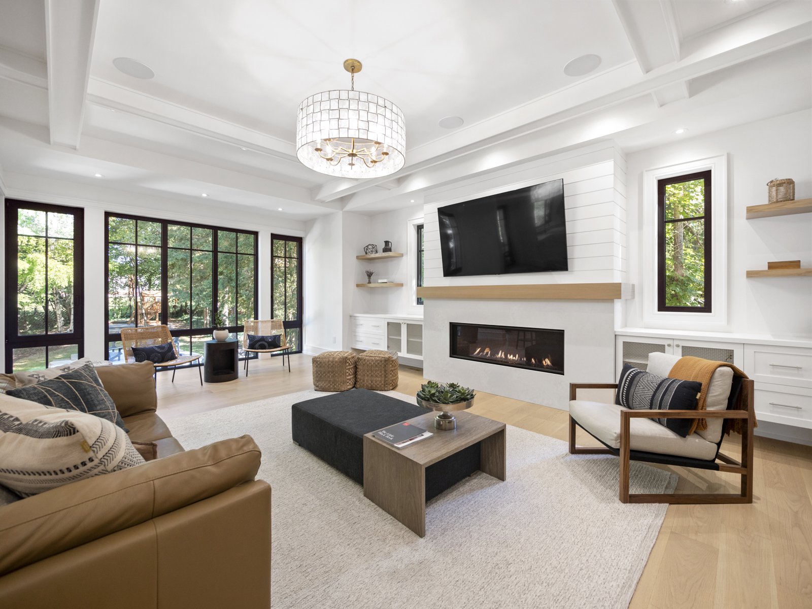 Modern living room with large windows, white walls, a fireplace, a large flat-screen TV mounted above the fireplace, a beige sofa, armchair, wooden coffee table with a small plant, and additional seating along the window wall.