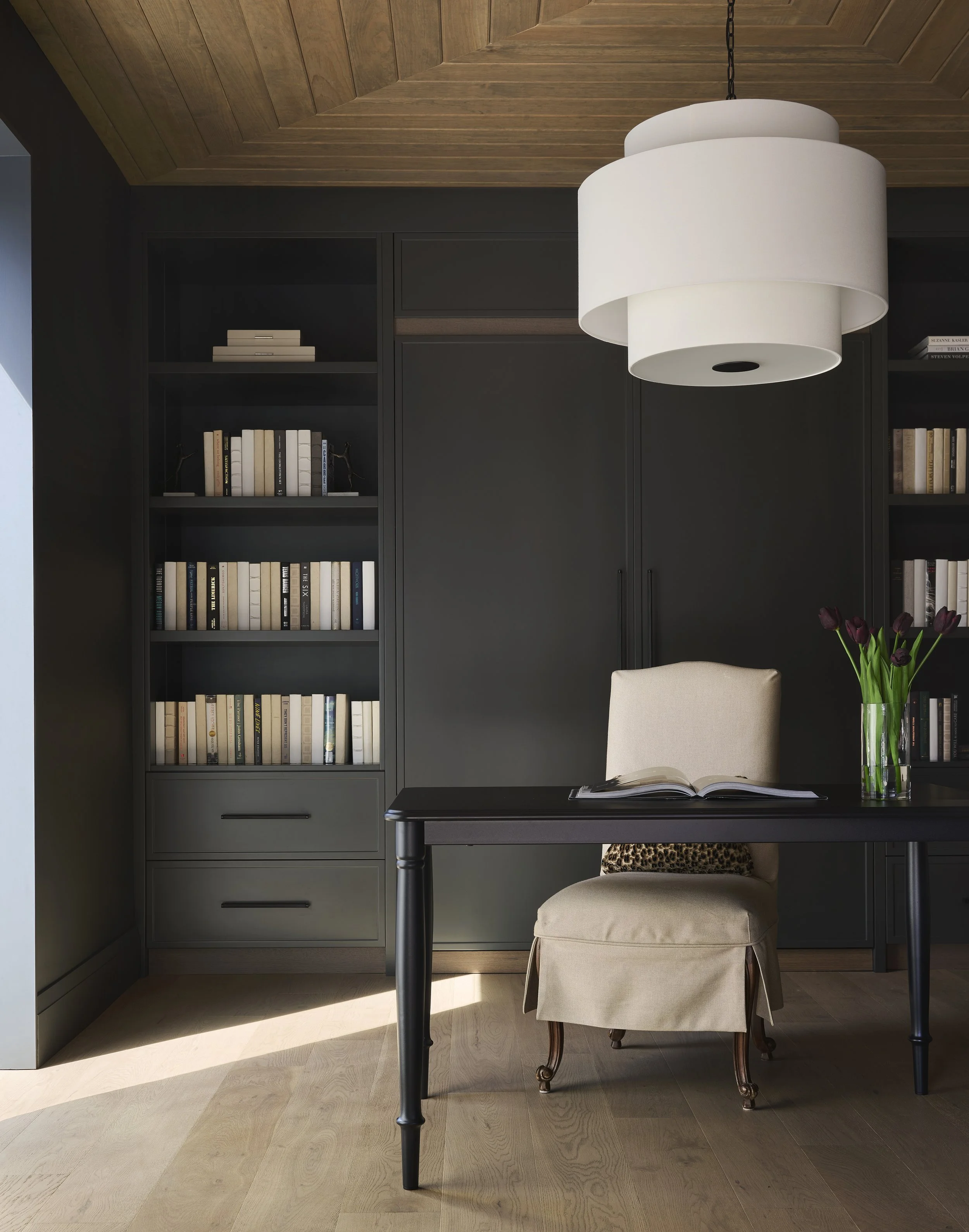 A home office space with a dark bookshelf filled with books, a beige upholstered chair with wooden legs, a black wooden desk, a clear glass vase with purple tulips, and a large white modern pendant light hanging from a wooden ceiling.