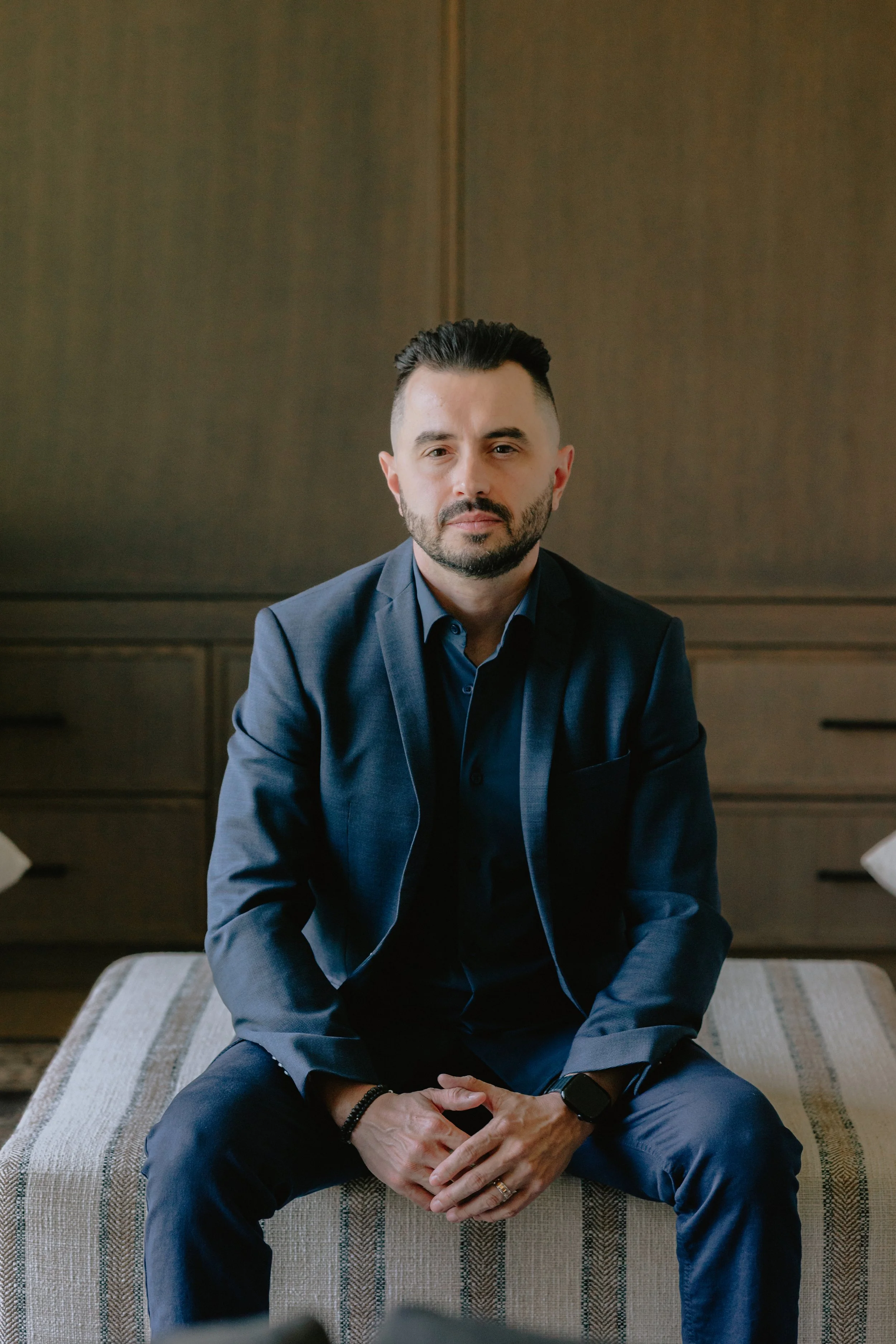 A man in a dark blue suit and shirt sitting on a striped upholstered bench in a wood-paneled room, looking directly at the camera.