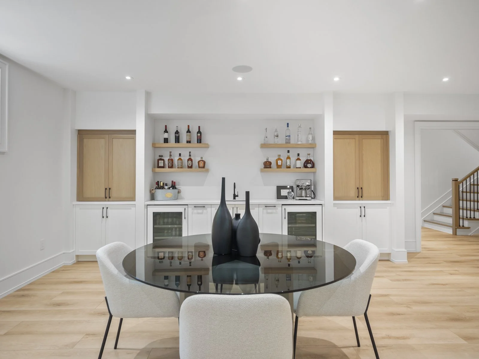 A modern dining area with a glass-top round table surrounded by four light-colored upholstered chairs, and a minimalist bar area with shelves of liquor bottles against a white wall.