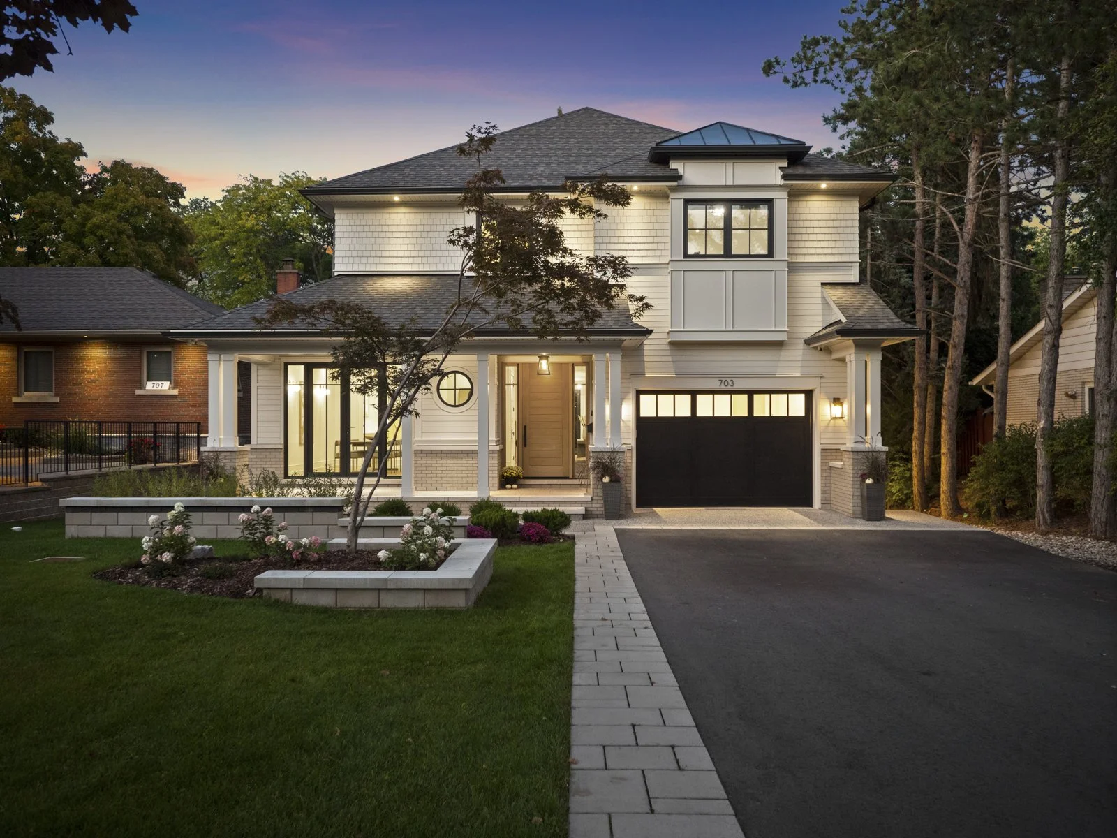 A modern two-story custom built house with a well-maintained front yard, black garage door, and exterior lighting at dusk.