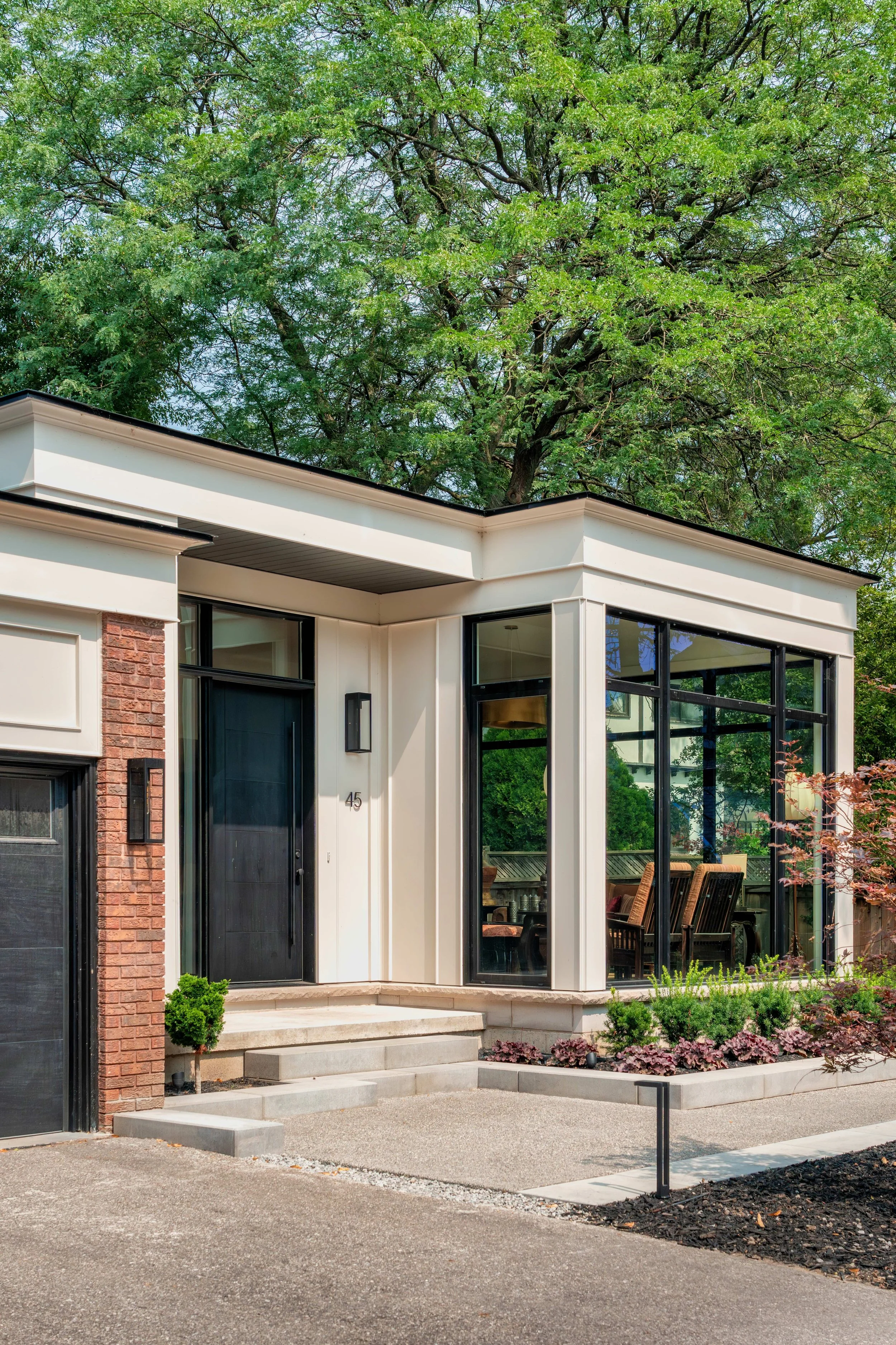 Modern house with a custom addition featuring large glass window, front steps, and landscaping, surrounded by tall trees.