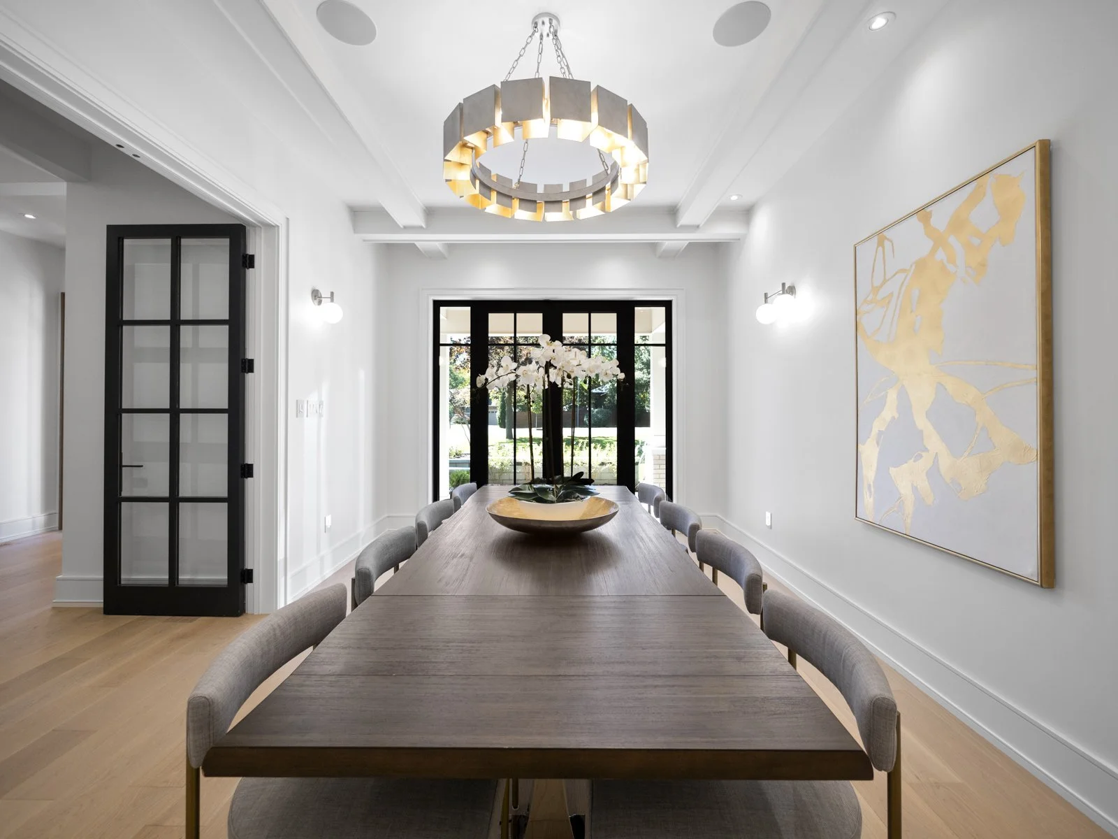 Elegant dining room with white walls, modern chandelier, large wooden table, gray chairs, a decorative bowl with white flowers, glass double front doors, and a large abstract artwork on the wall.