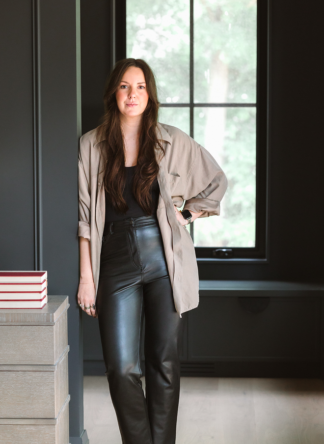 A young woman with long brown hair is standing indoors next to a gray piece of furniture with a stack of books on it. She is wearing a black top, black leather pants, and an oversized beige blazer. There is a window behind her revealing green trees outside.