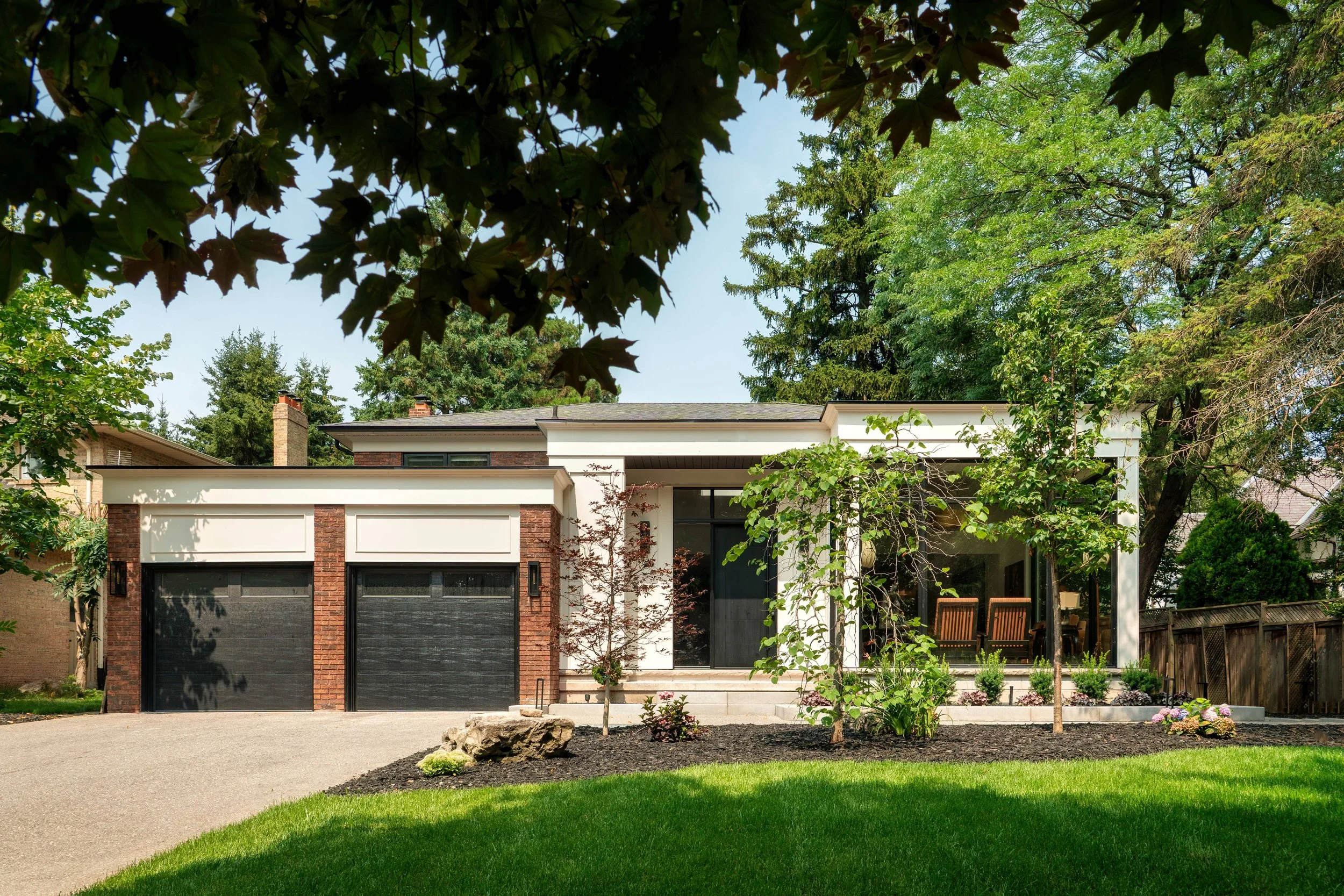 Modern house with two black garage doors, brick accents, large windows, a manicured lawn, trees, and plants in the front yard, surrounded by greenery.